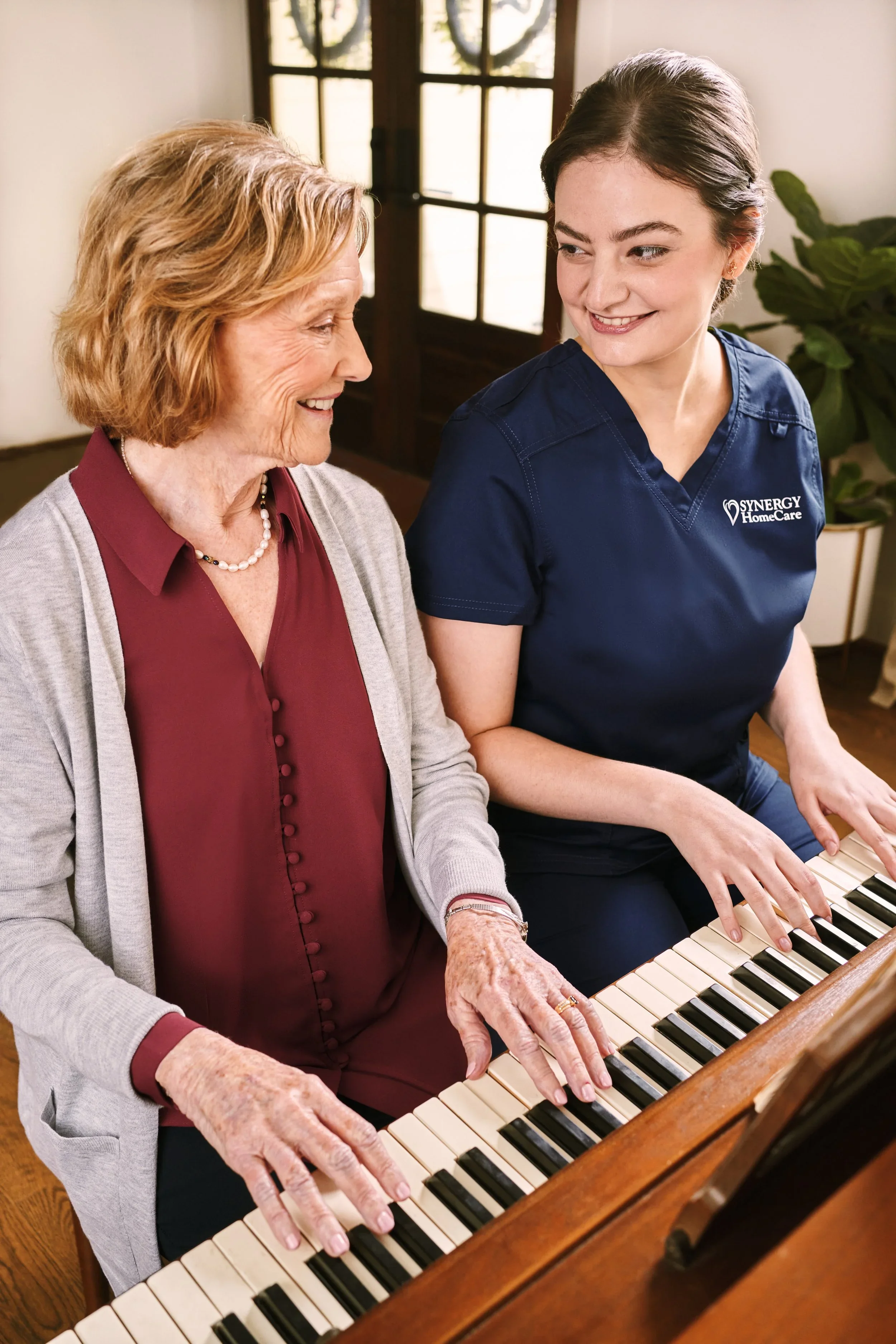 Elderly woman and young woman playing piano together indoors, smiling at each other.