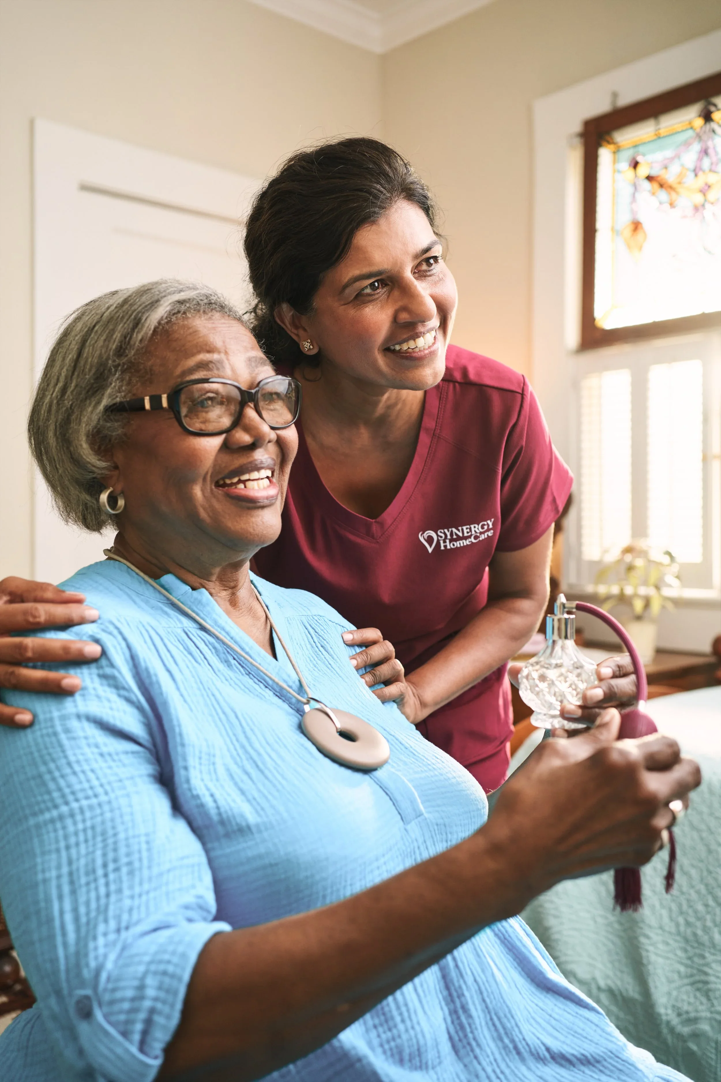 A healthcare worker in maroon scrubs smiling and standing next to an elderly woman with glasses, gray hair, and a blue medical gown, who is holding a glass likely connected to a medical device. They are in a room with sunlight, a window with stained 