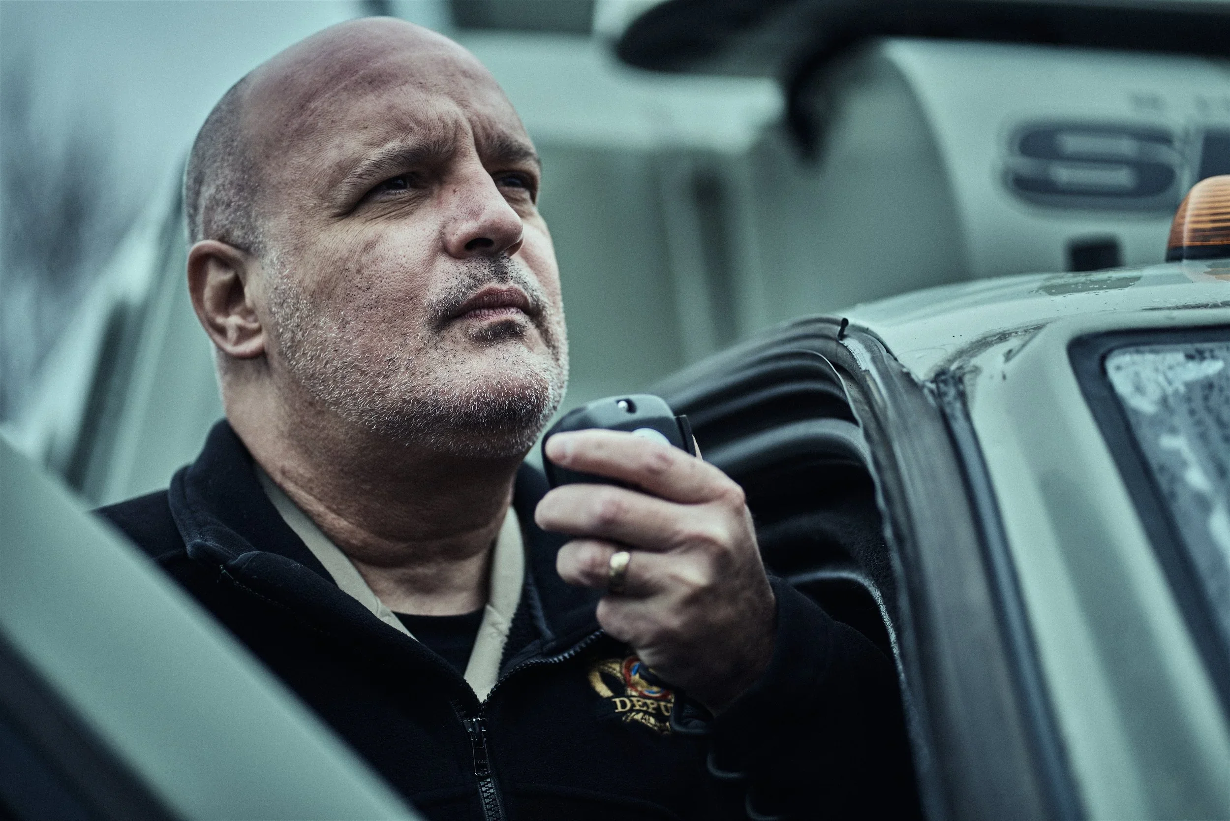 A man with a serious expression, short, bald head, and stubble on his face, wearing a black jacket and holding a walkie-talkie, is looking out from inside a vehicle.
