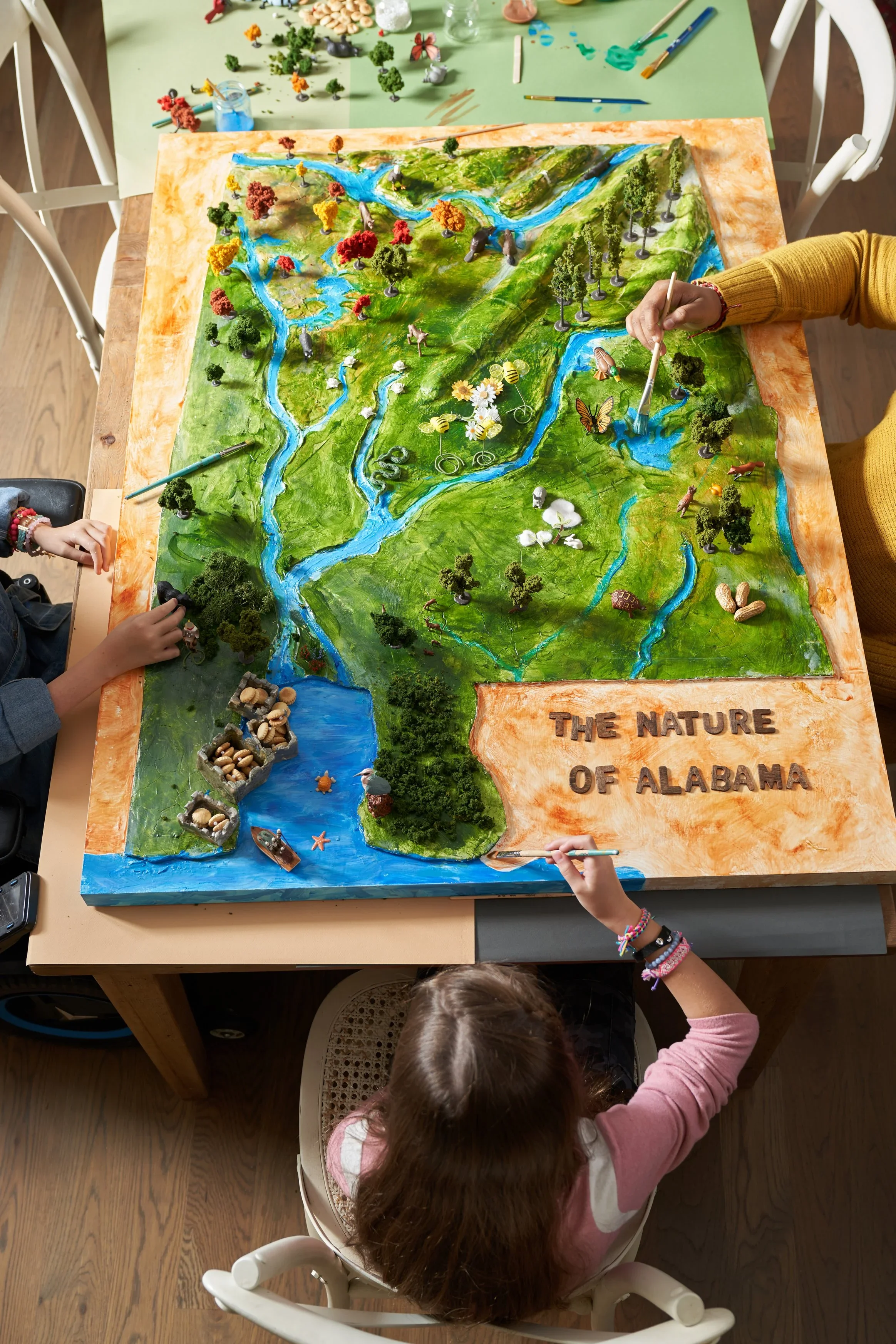 Children working on a detailed 3D map of Alabama showing rivers, trees, and wildlife, with the title 'The Nature of Alabama' on the sheet.
