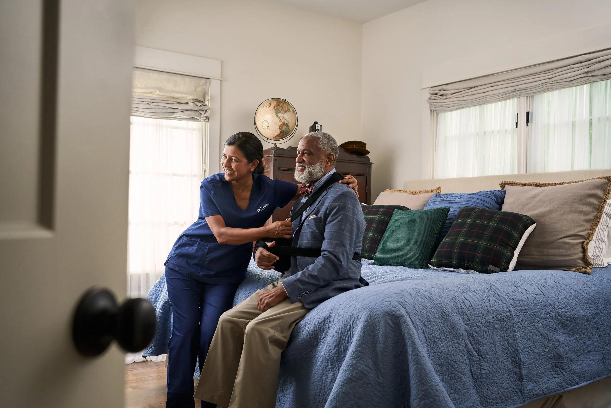 A nurse helping an elderly man with a medical device in a bedroom with a bed and pillows, sunlight through the windows, and a world globe on a cabinet.