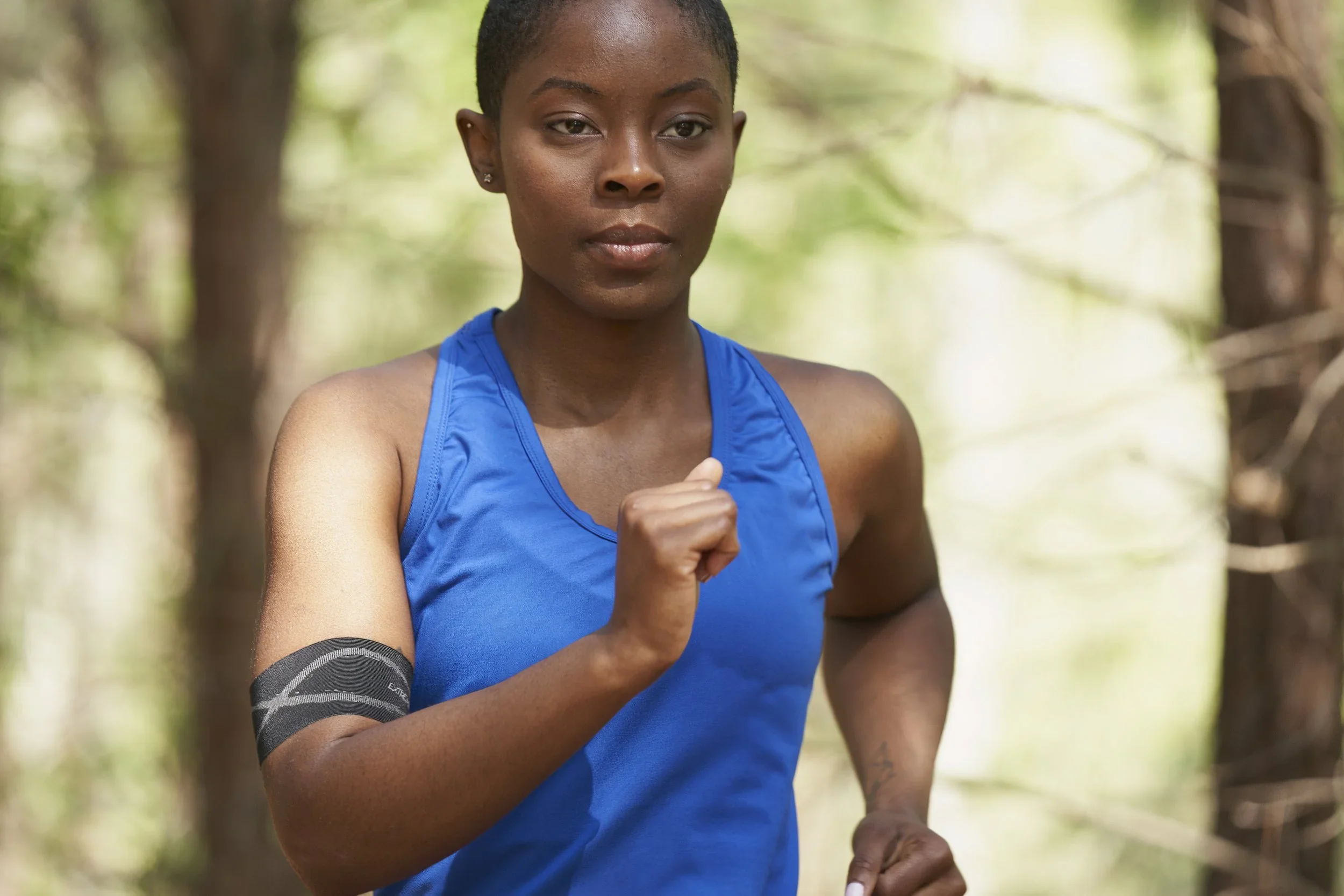 A young woman with short hair wearing a blue tank top running outdoors in a green, wooded area.