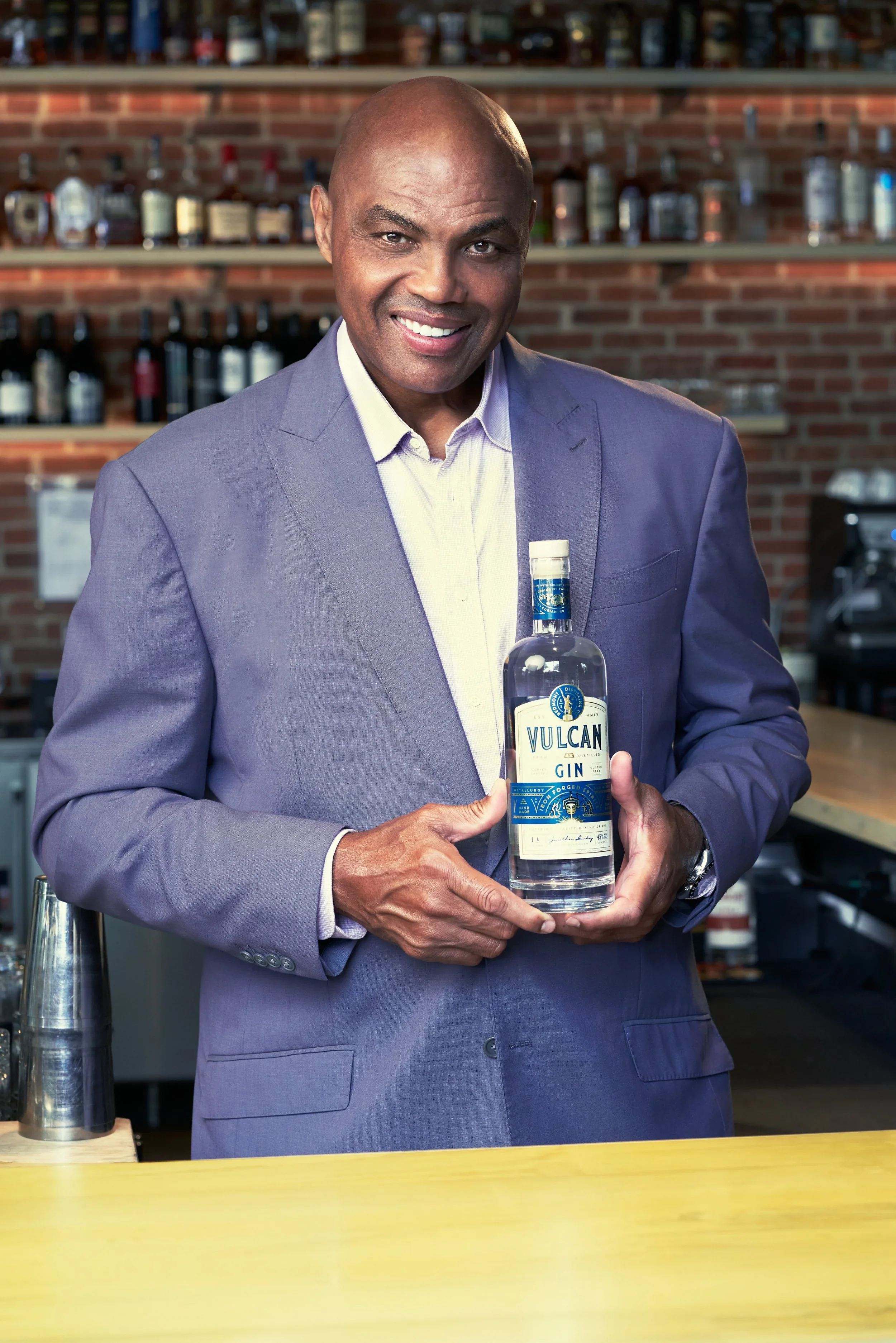 A middle-aged man in a gray suit holds a bottle of Vulcan gin in a bar with shelves of liquor bottles in the background.