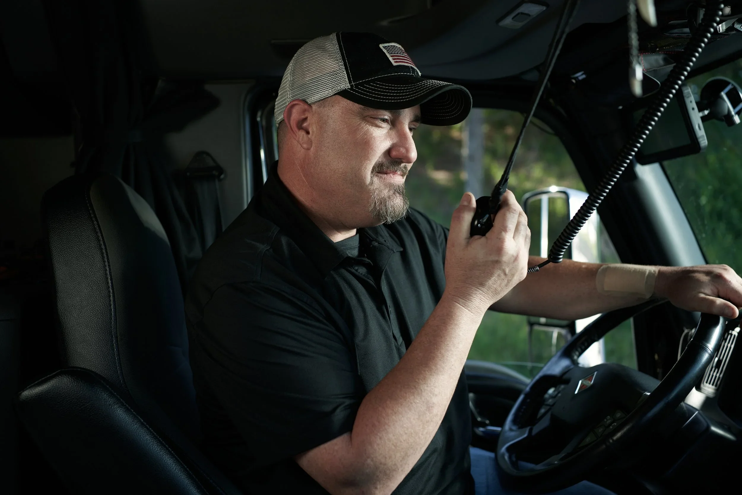 A man wearing a trucker hat with a USA patch, sitting inside a vehicle, speaking into a radio microphone.