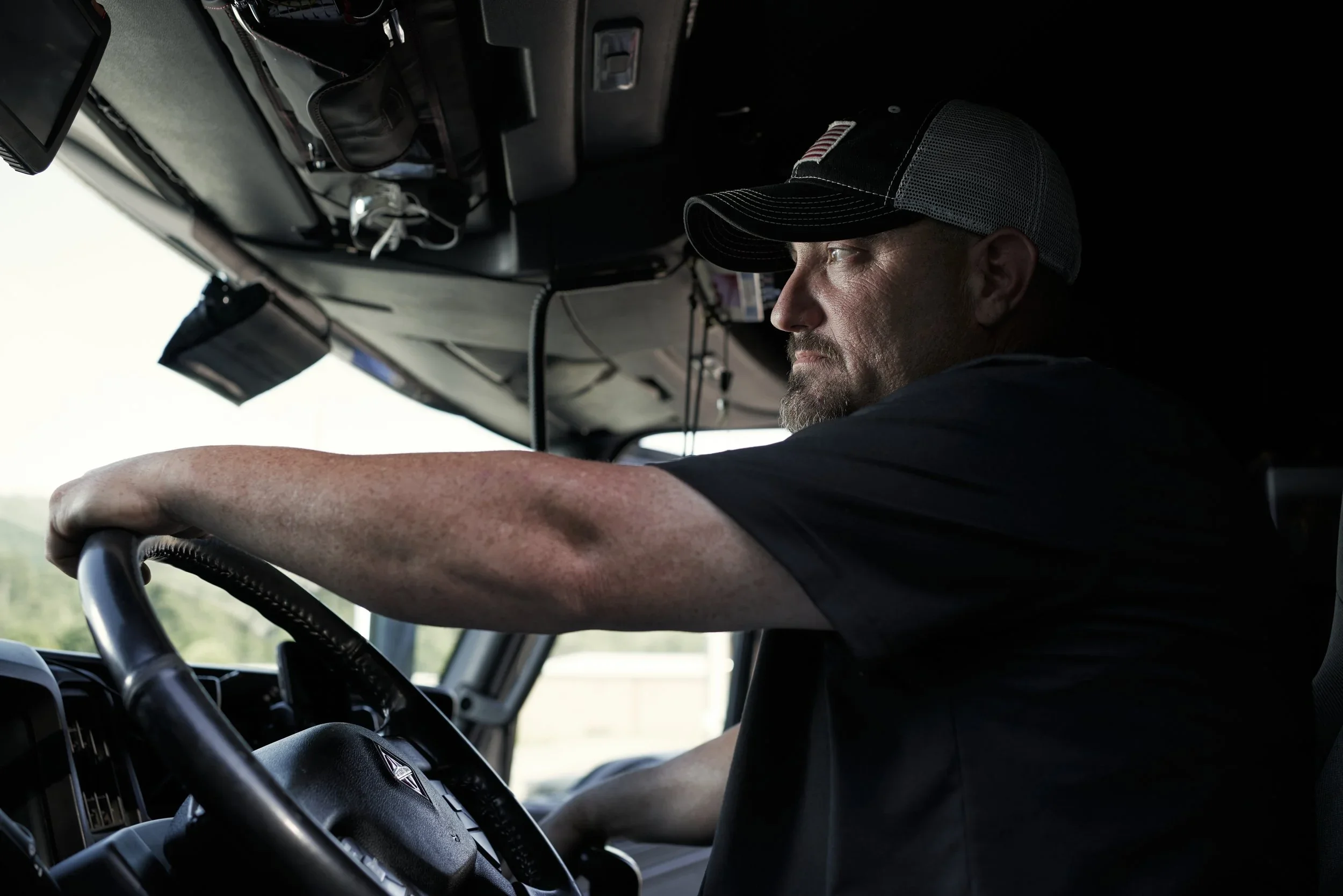A man with a beard and a trucker cap driving a vehicle, visible from the side with his arm stretching out towards the steering wheel.