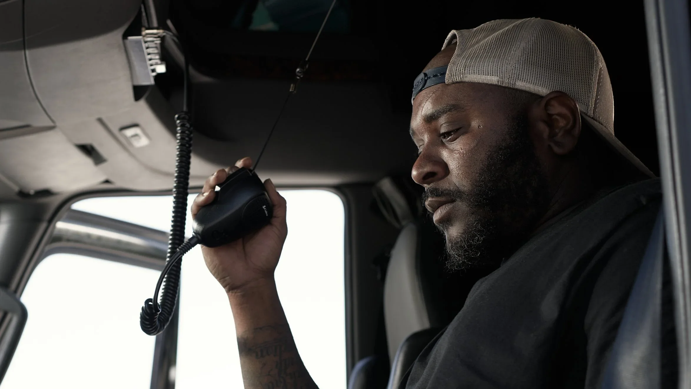 A man with a beard and wearing a beige baseball cap uses a radio microphone while seated inside a vehicle, possibly a truck.