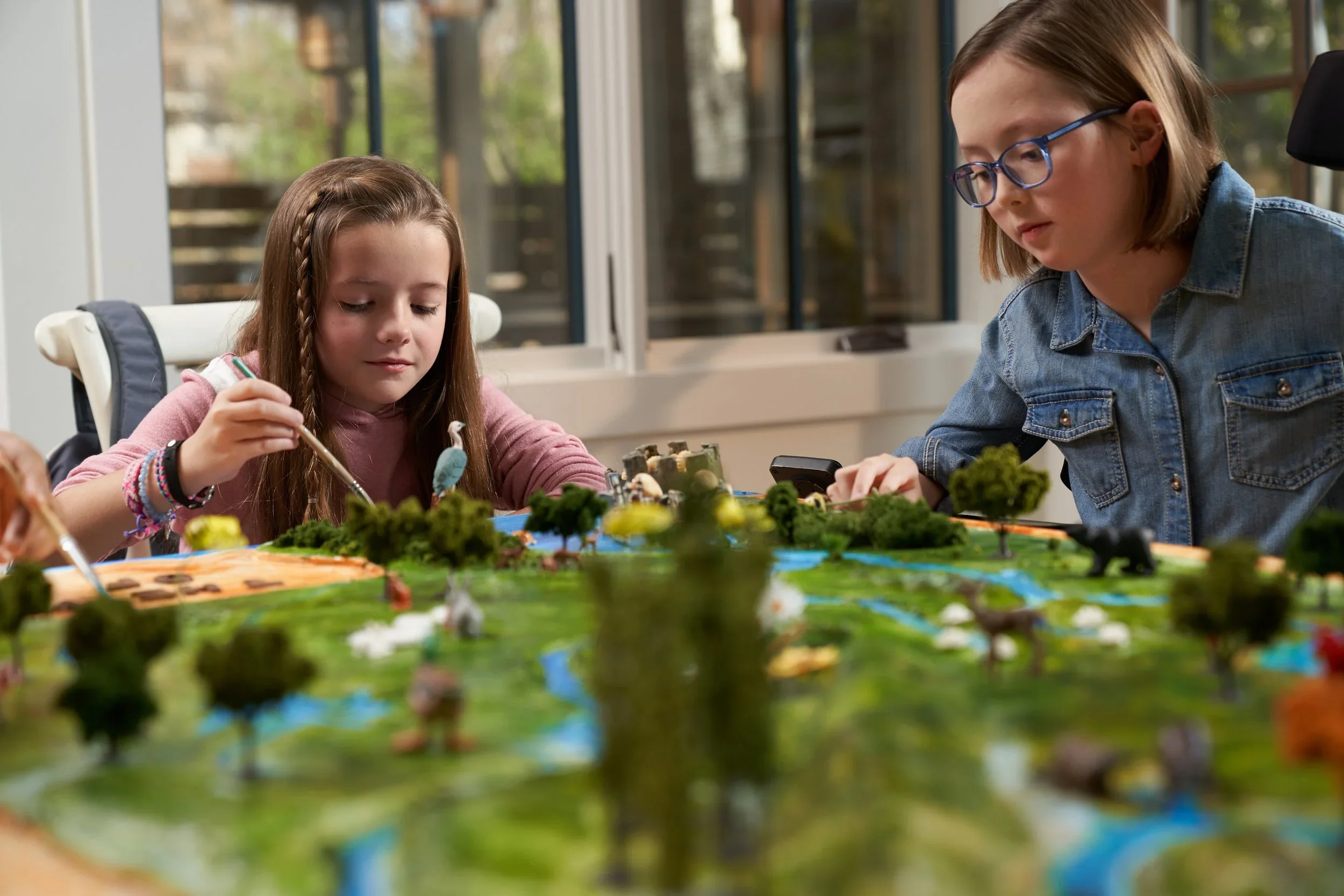Two young girls working on a detailed miniature landscape model with trees and water features, seated at a table near large windows.