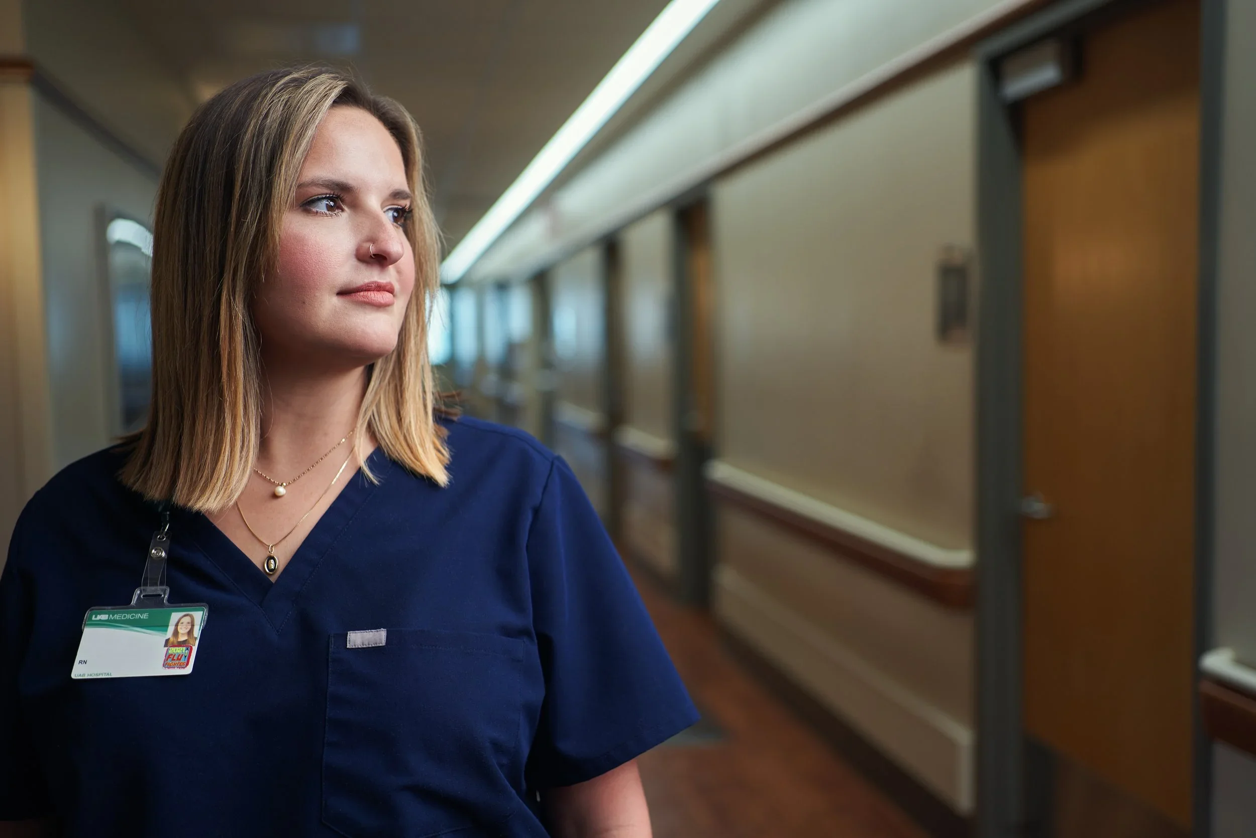 A female nurse in navy scrubs with a hospital ID badge stands in a hospital corridor.