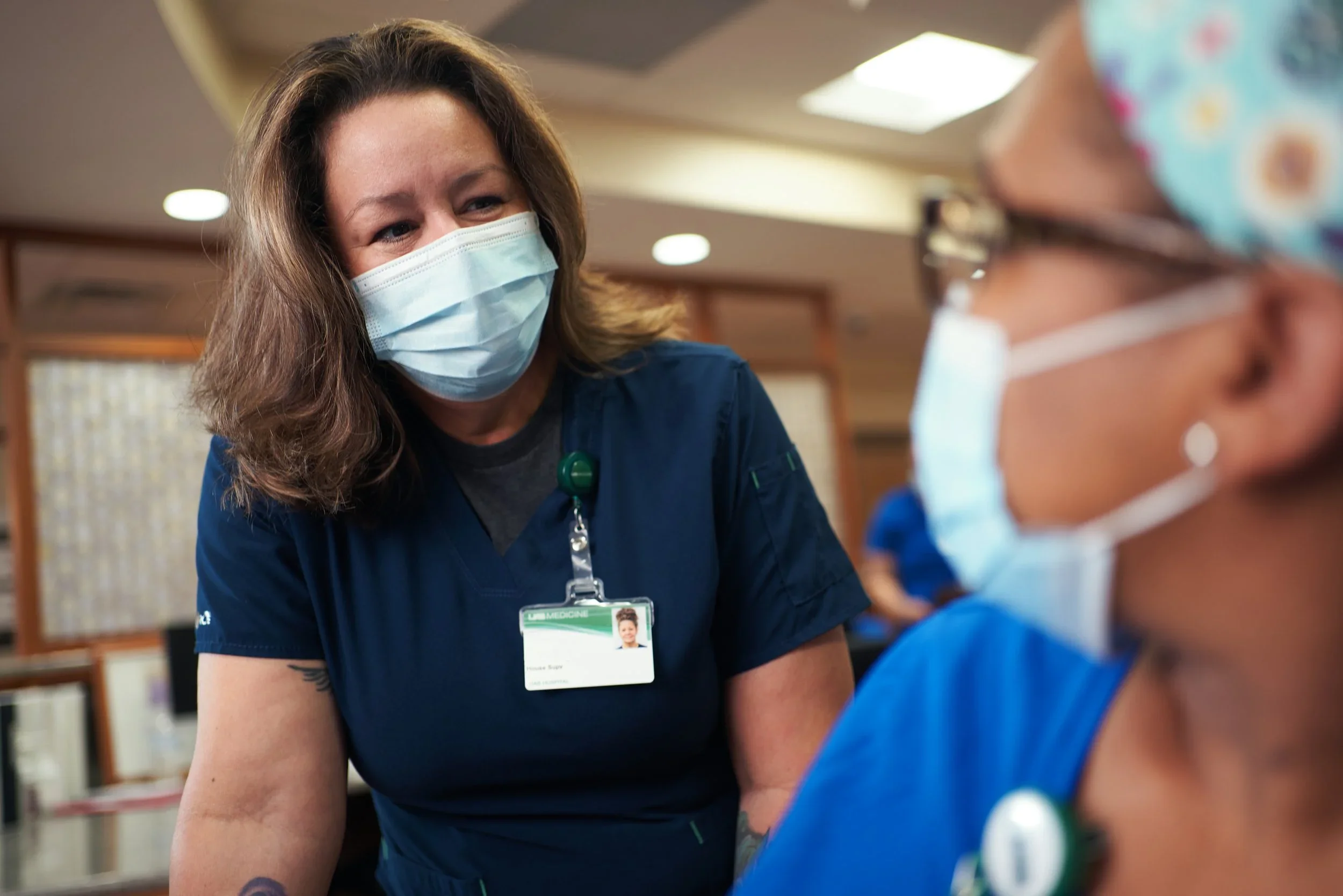A healthcare worker, wearing a face mask, hair down, and an ID badge, is smiling and looking at a young patient in a medical setting.
