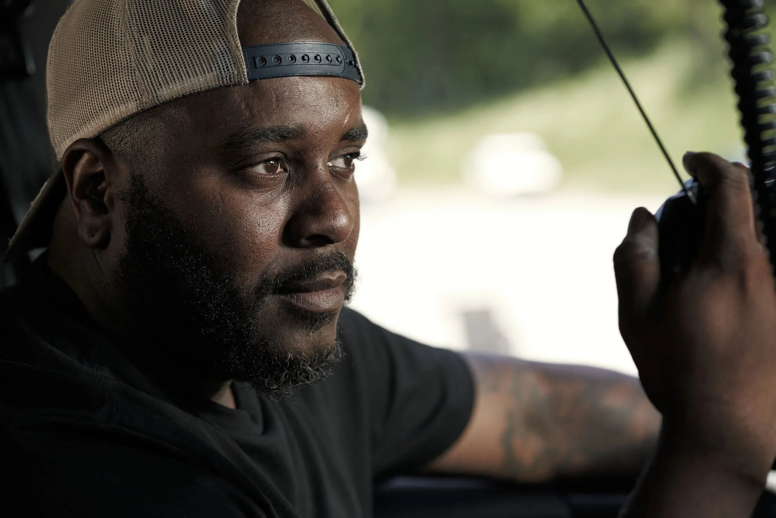 A close-up of a man with a beard wearing a beige cap backwards, focusing intently on a walkie-talkie he is holding.