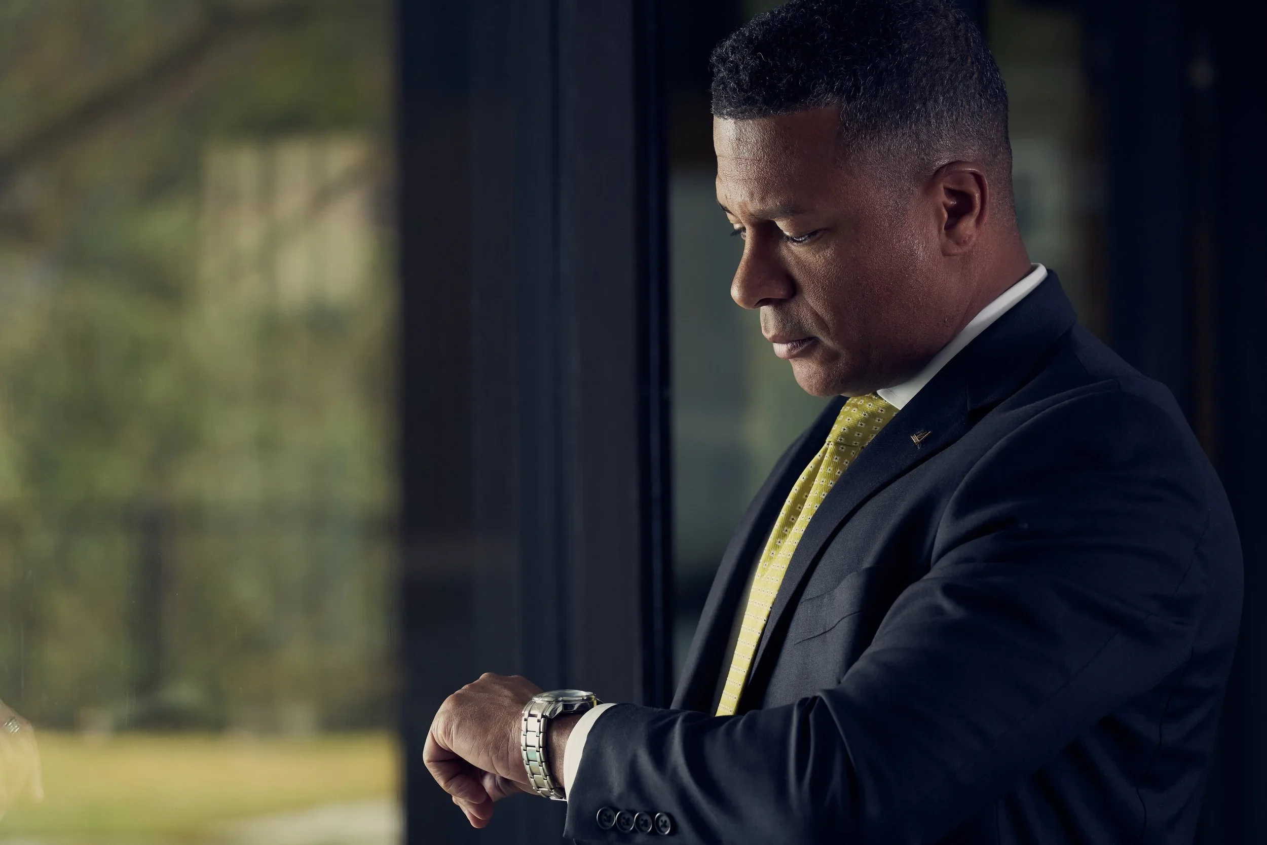 A man in a dark suit with a yellow tie looking at his wristwatch, standing indoors near a window.