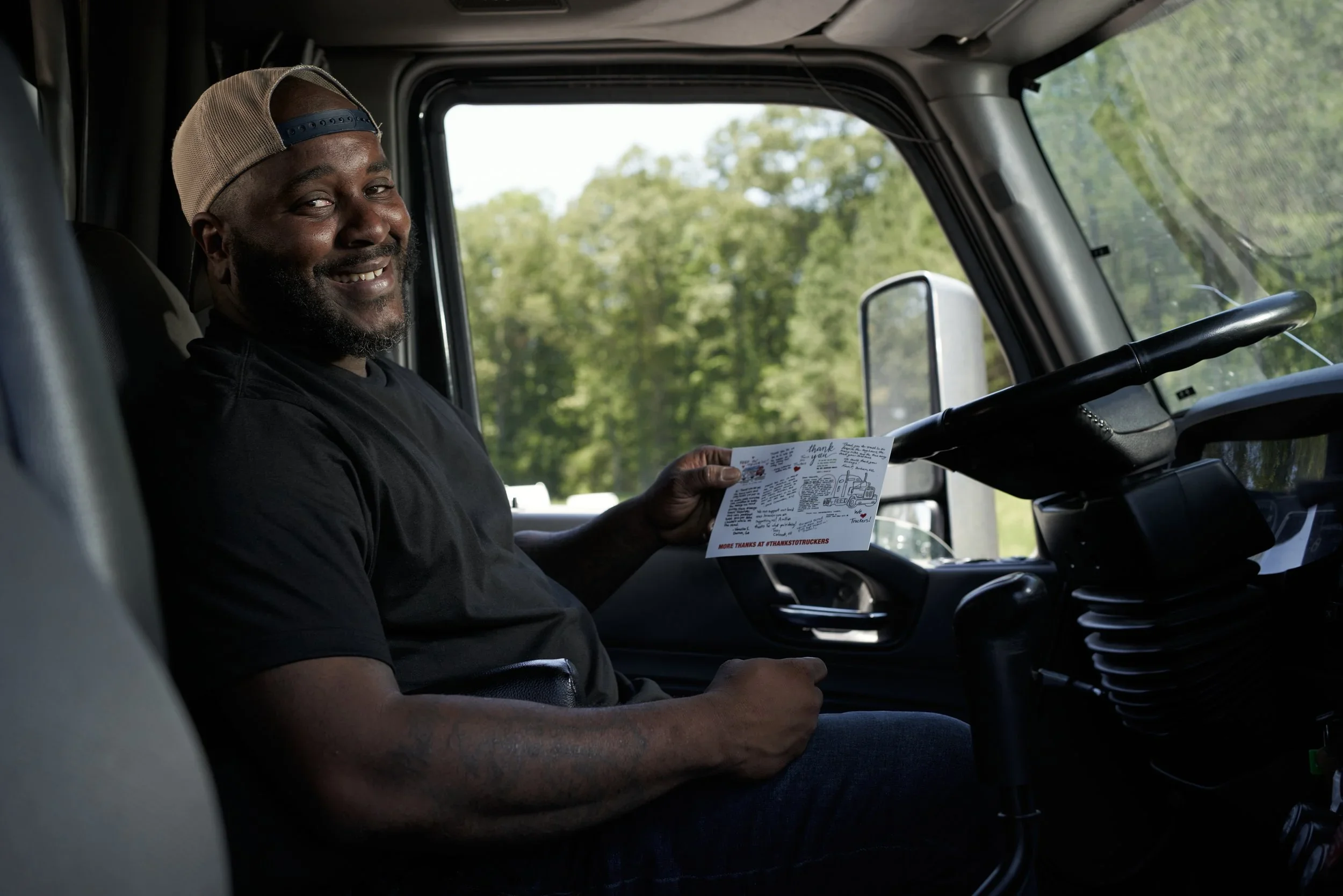 A smiling man sitting inside a truck, holding a thank you card, with green trees visible through the truck window.