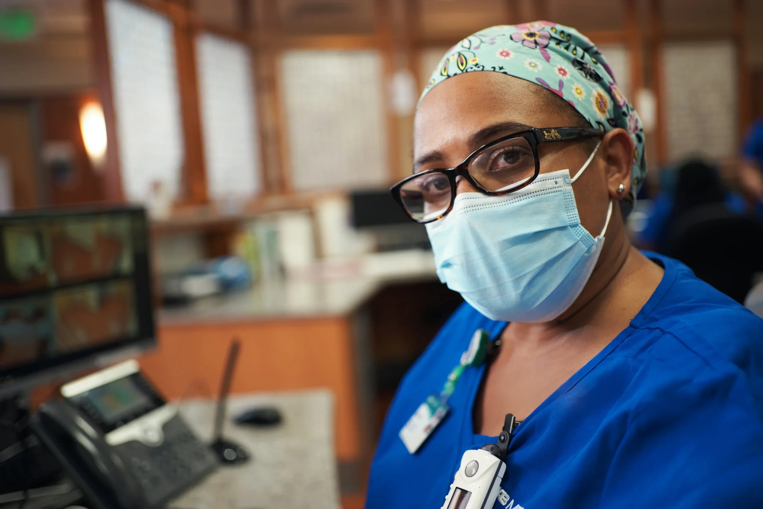 A healthcare worker wearing glasses, a colorful headscarf, a face mask, and blue scrubs, sitting at a desk with a computer and medical devices, in a hospital or clinical setting.