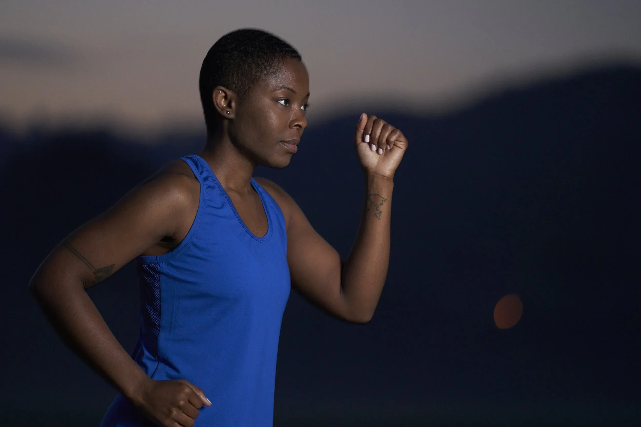 A woman with short hair wearing a blue athletic tank top, lifting her right arm in a fist, against a dark background.