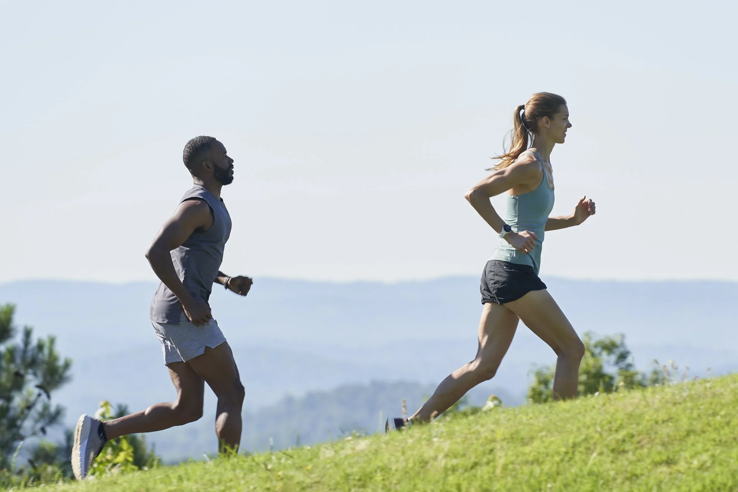 Two people running outdoors on grassy land with a scenic view and blue sky.