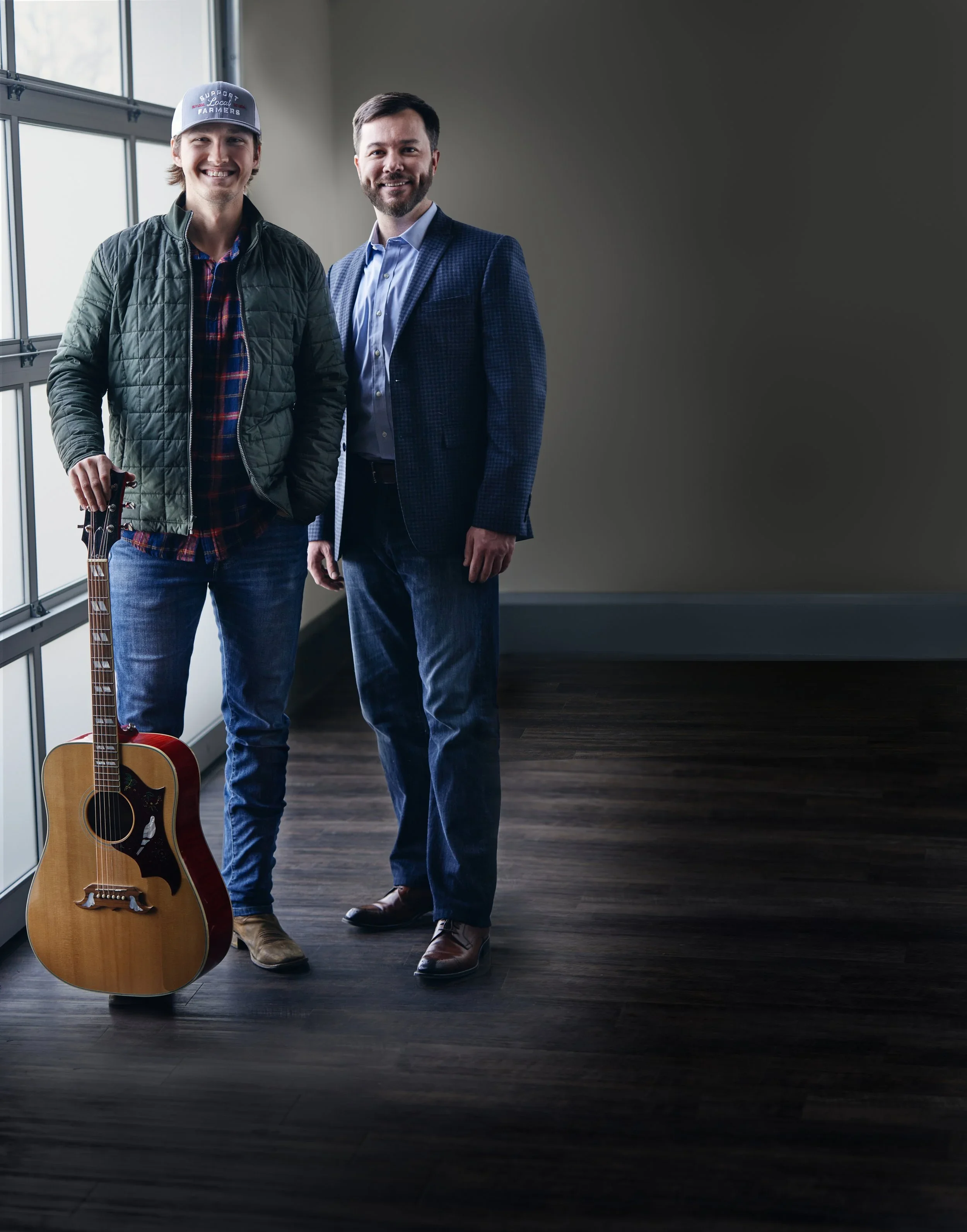 Two men standing indoors next to a large window. One man is holding a guitar, and both are smiling. One is wearing a blue checked blazer, and the other is wearing a hat, a quilted jacket, and a plaid shirt.