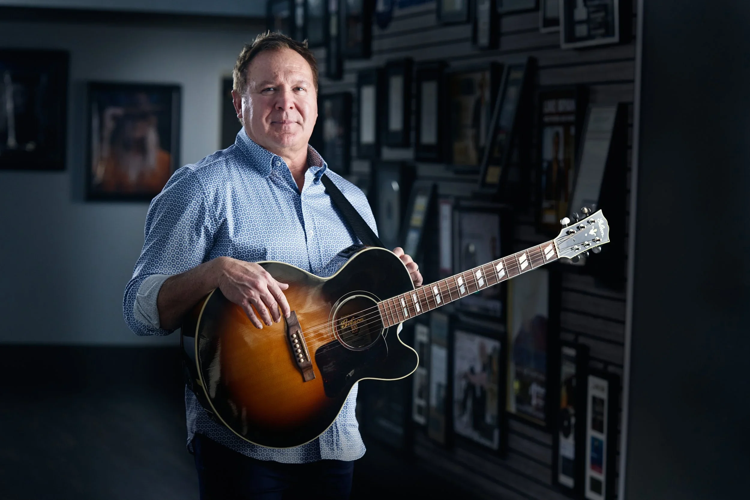 A man with short brown hair wearing a blue patterned button-up shirt playing an acoustic guitar in a room with framed pictures or artwork on the wall.