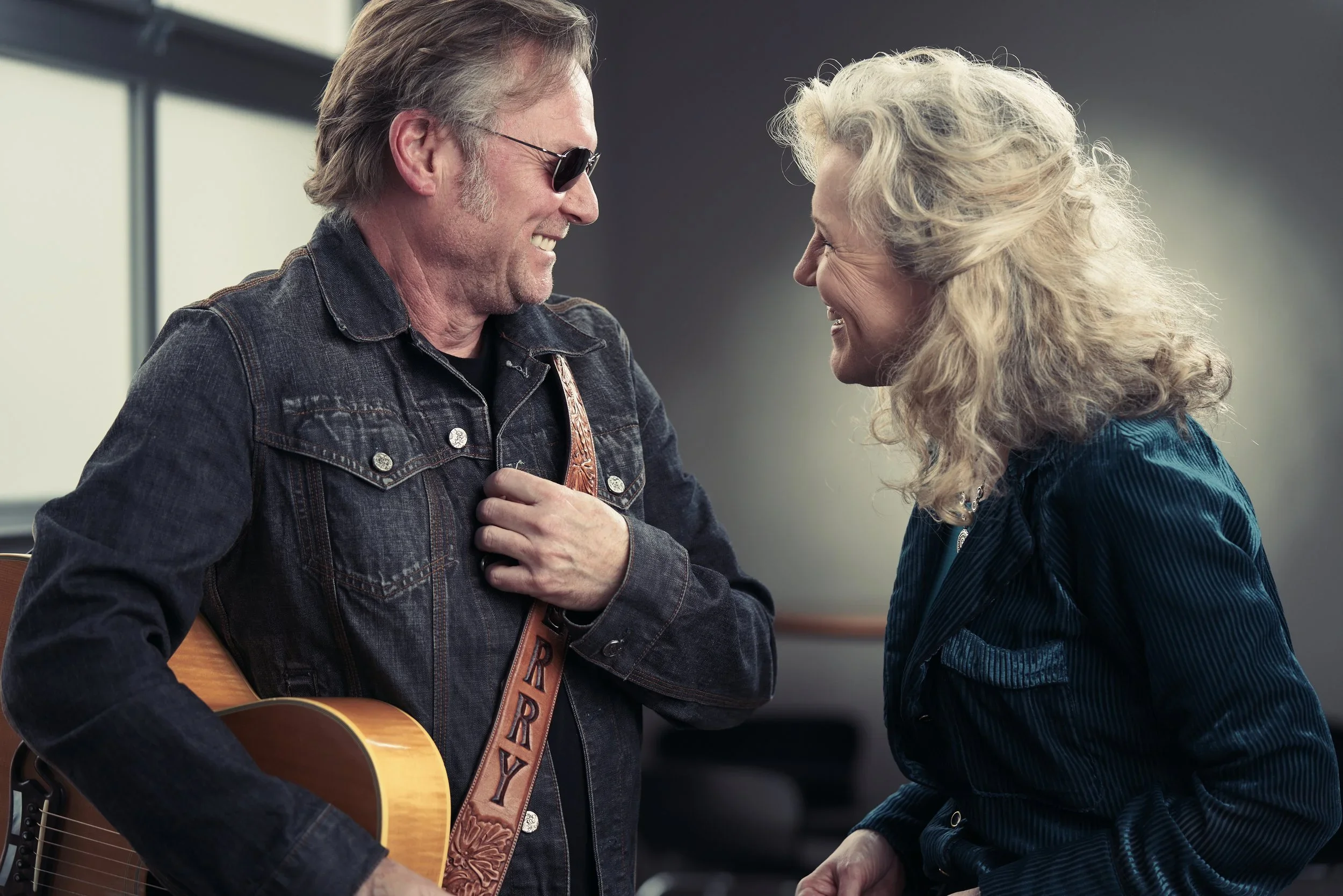 A man wearing sunglasses and a denim jacket smiling in front of a woman with curly blonde hair, who is also smiling, as they face each other closely in a room.