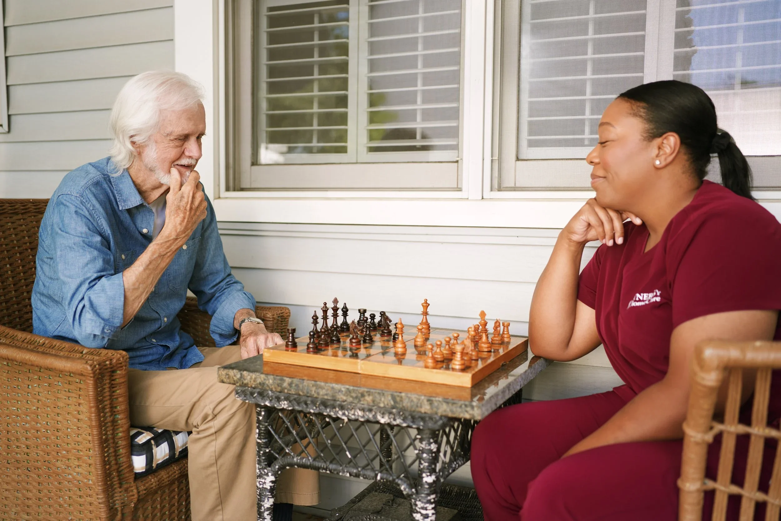 An elderly man and a young woman playing chess on a porch.