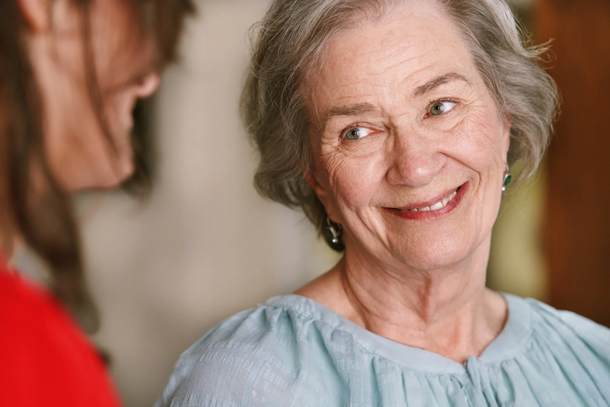An elderly woman with gray hair and green eyes smiling while talking to a woman with long brown hair, who is out of focus.