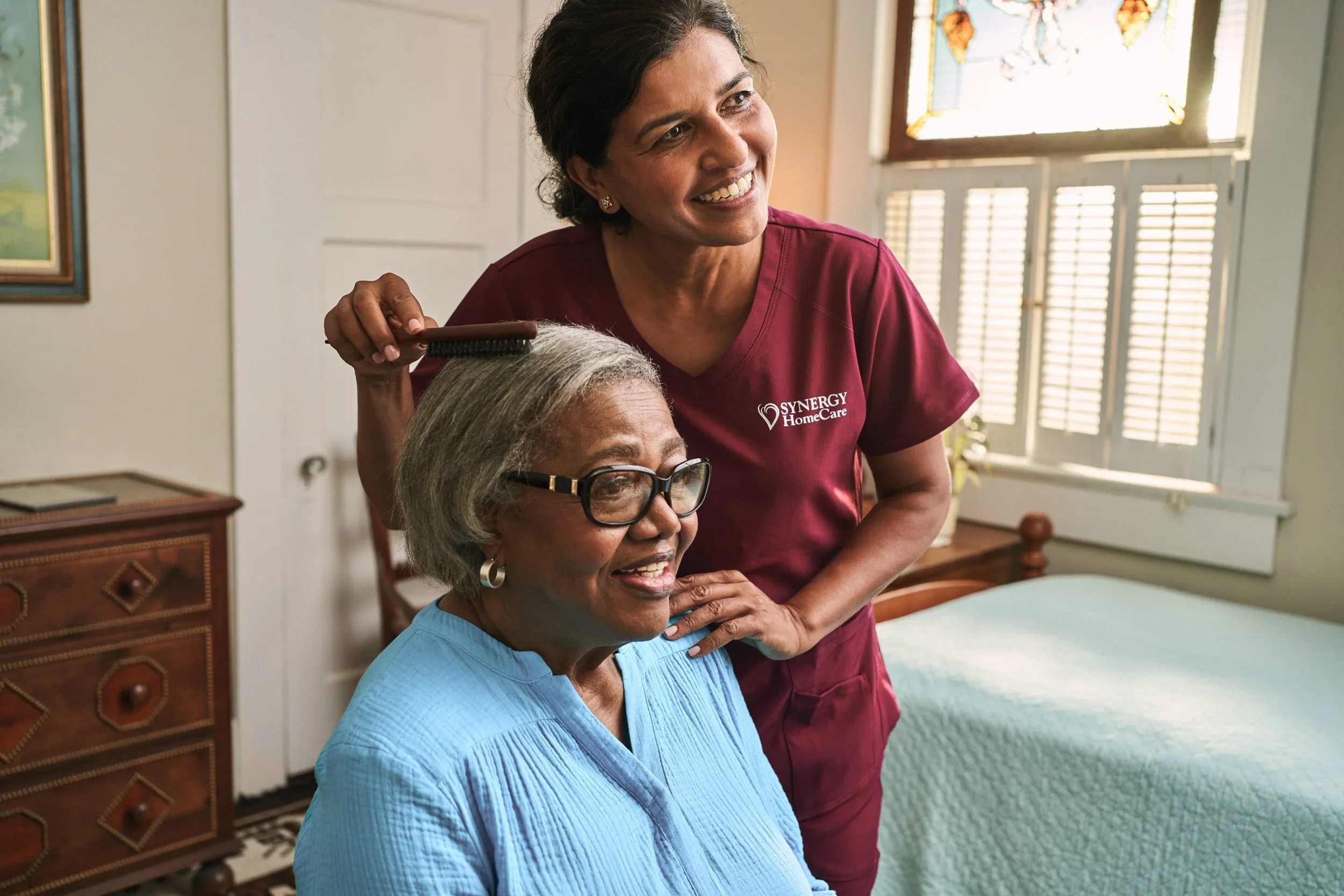 A caregiver brushing the hair of an elderly woman sitting in a bedroom.