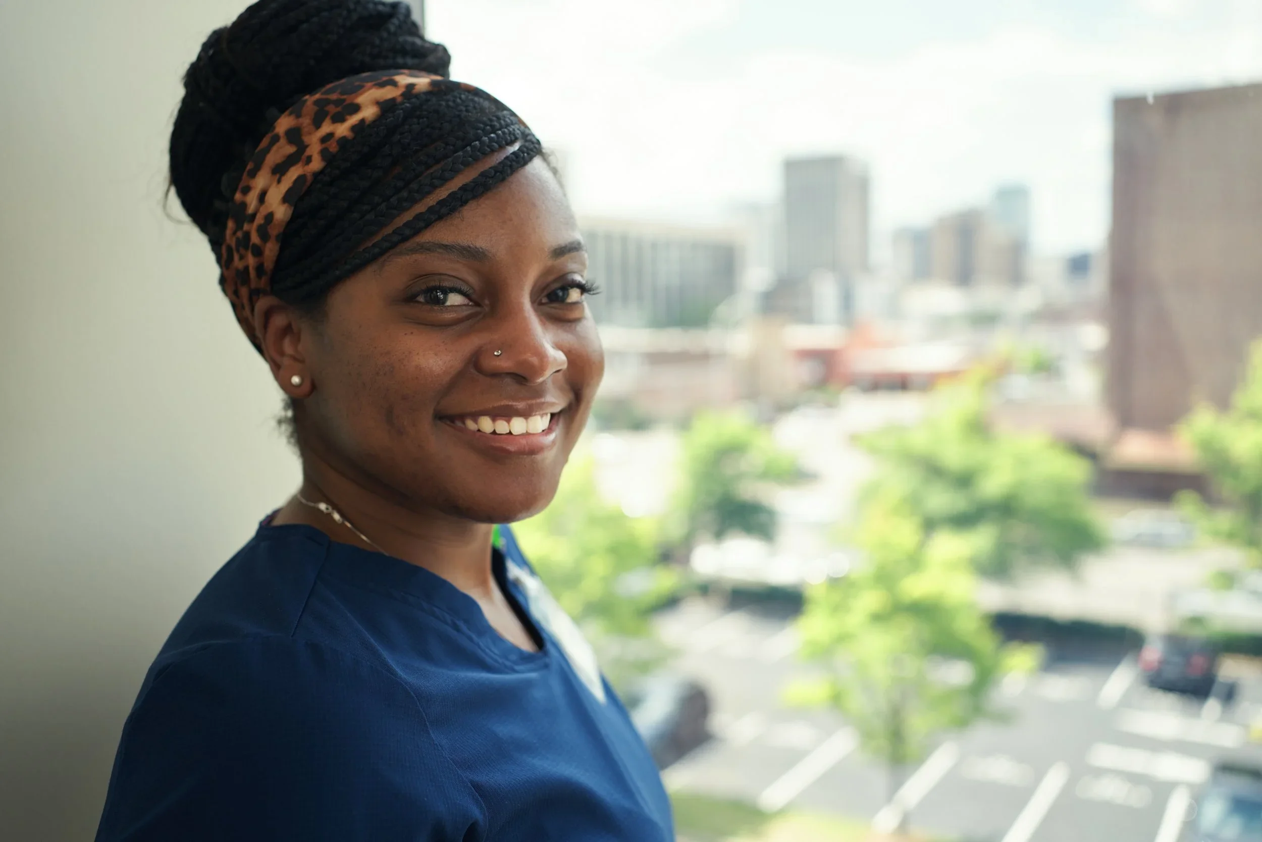 A woman with a braid hairstyle, wearing a leopard-print headband and pearl earrings, smiling and looking at the camera. She is dressed in a navy blue top, standing near a window with a cityscape in the background.