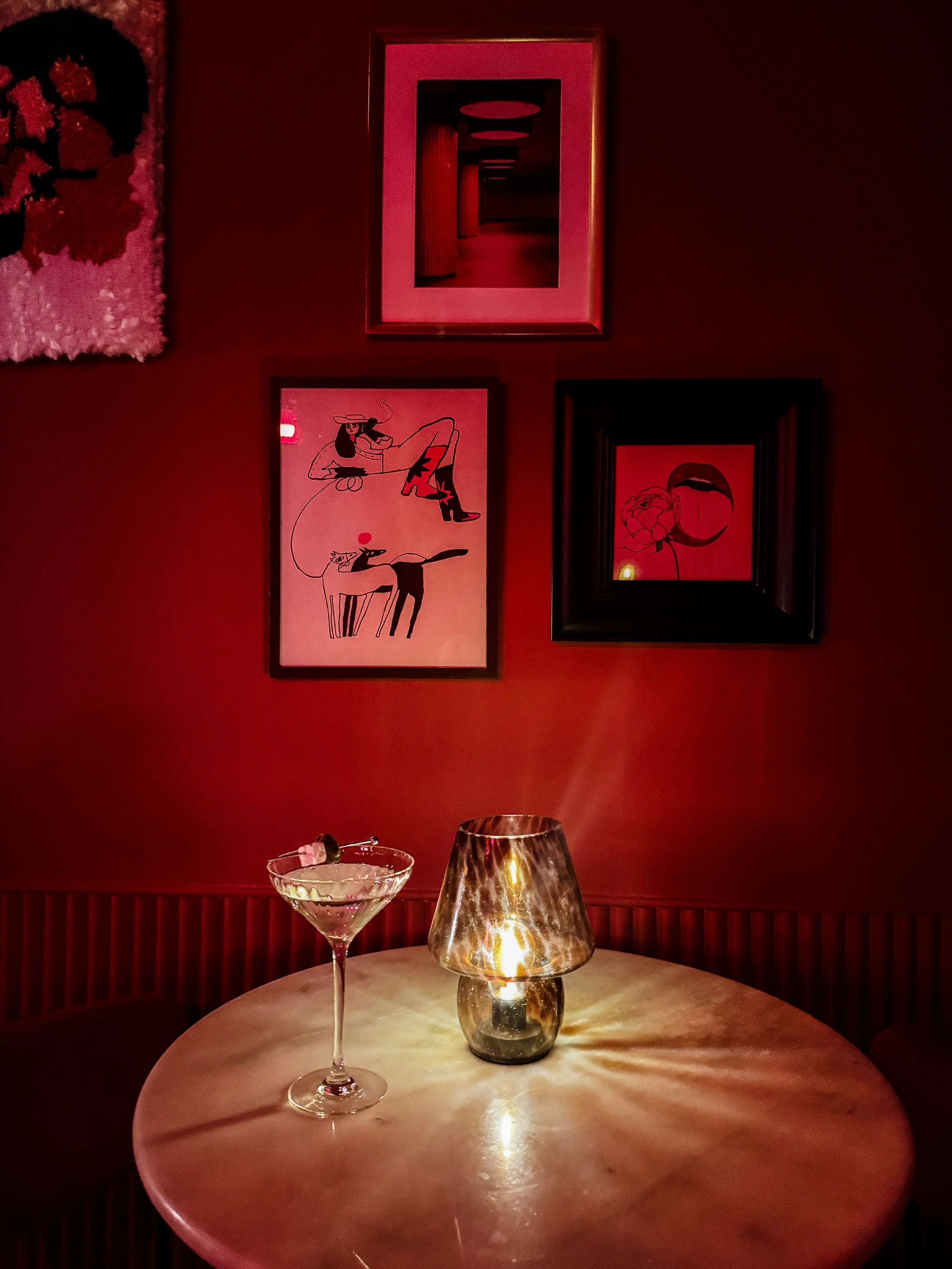 A round table with a martini glass, a small candle, and a lava lamp in a dimly lit room with red walls, framed artwork, and candles.