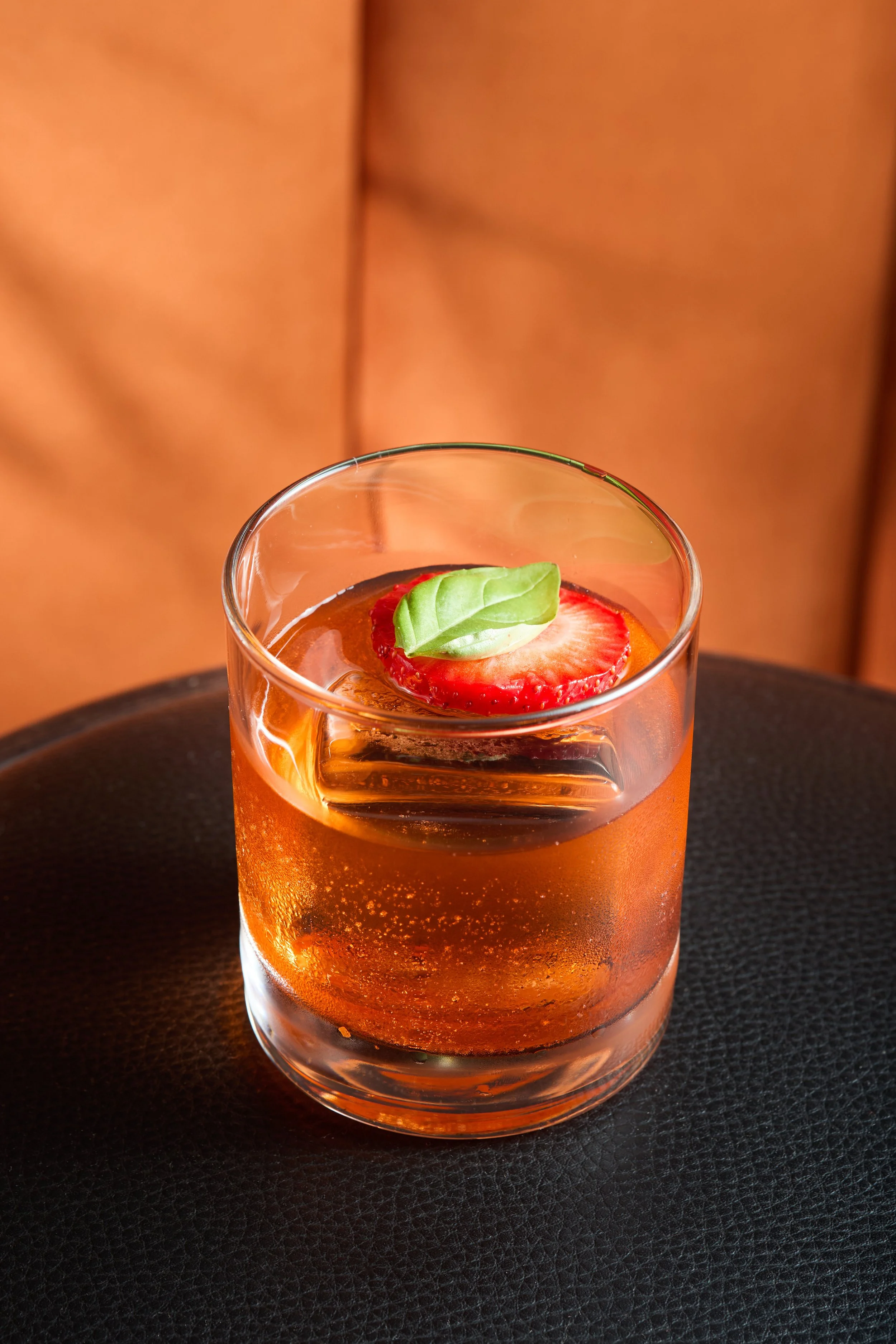 A glass of sparkling beverage topped with a slice of strawberry and a basil leaf, placed on a dark surface.