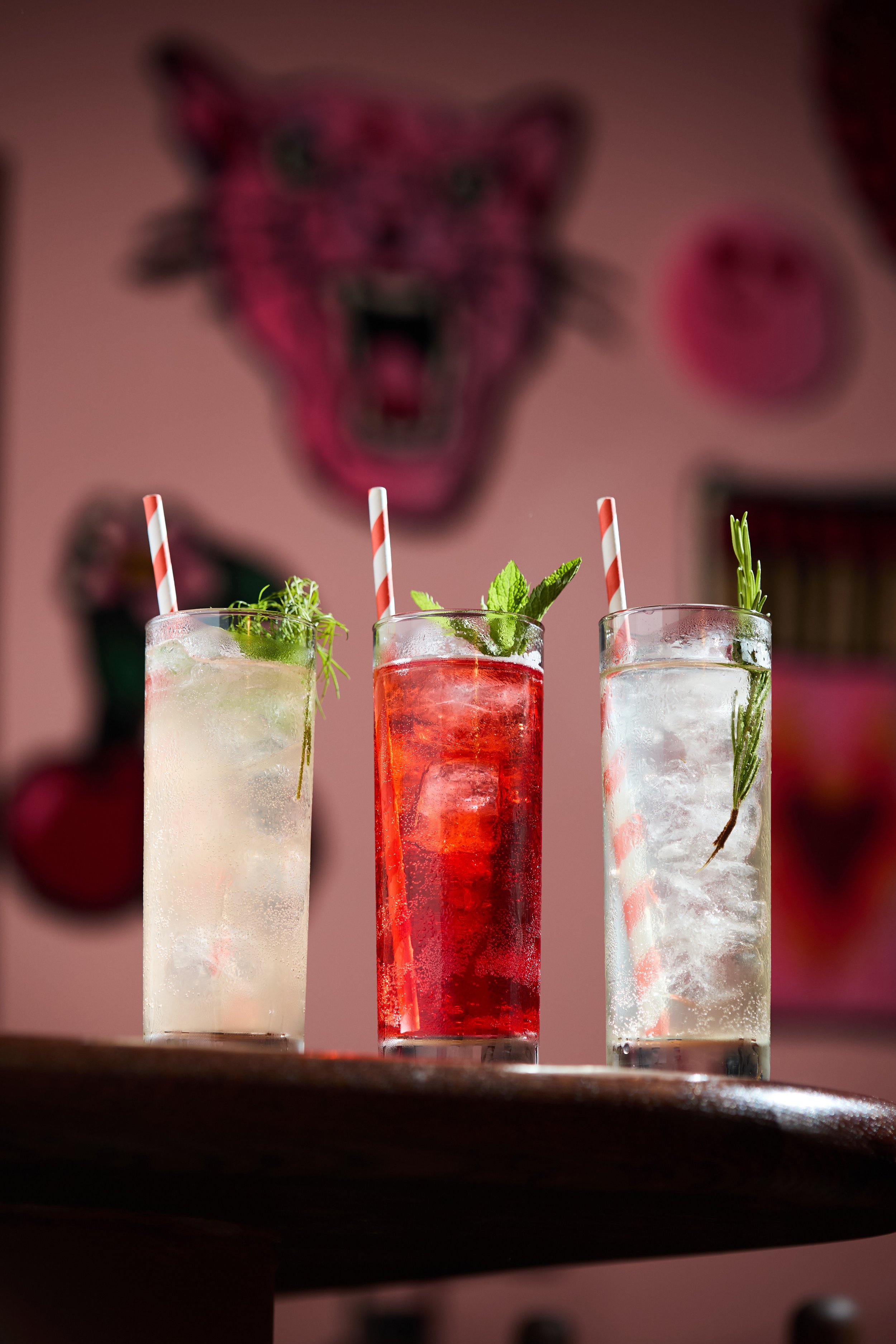 Three colorful cocktails with garnishes, garnished with mint, rosemary, and herbs, and striped straws, in front of a pink wall with artwork including a leopard head and face stickers.