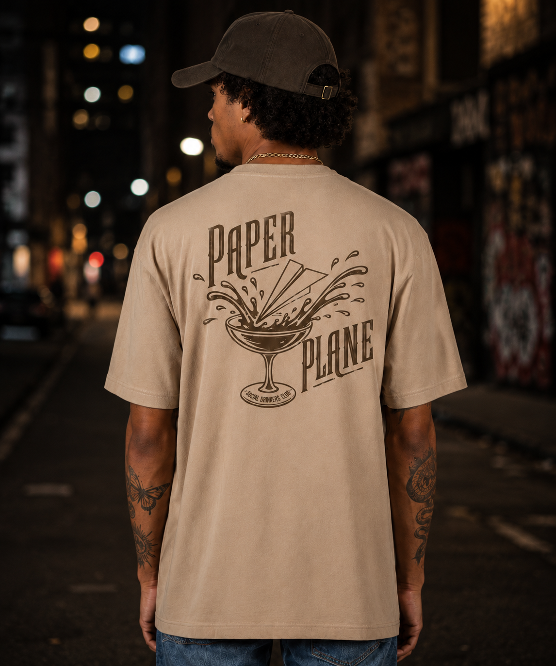 A man wearing a gray cap and beige T-shirt with a graphic and text on the back, standing in an urban setting at night with blurred streetlights and buildings in the background.