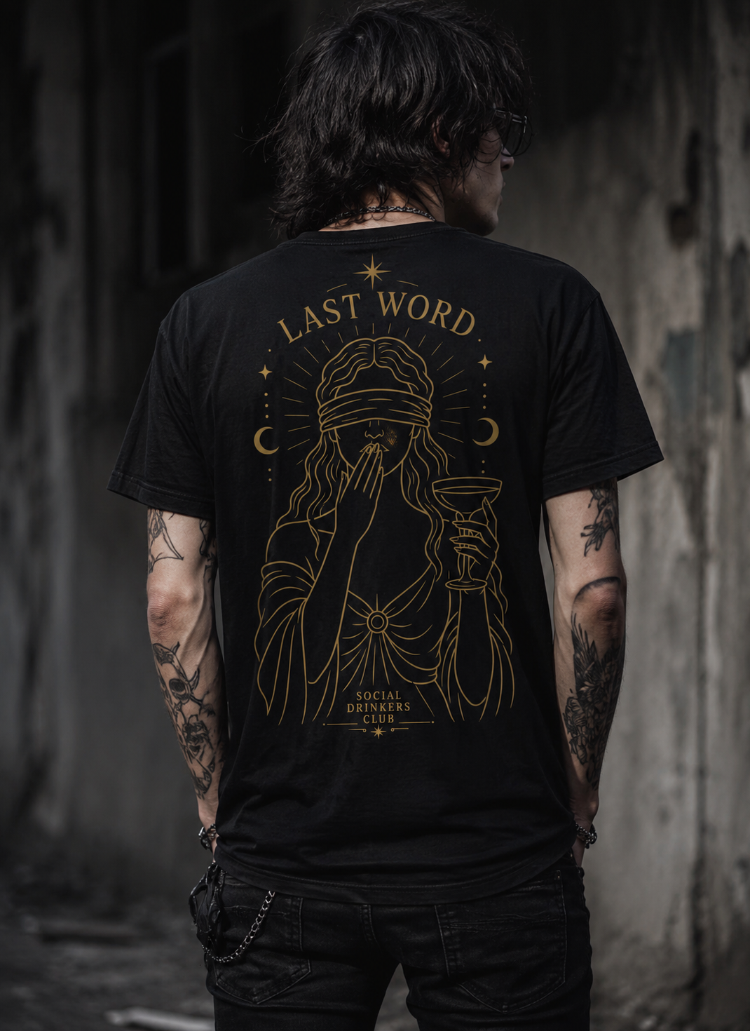 A person with tattoos on both arms, wearing a black T-shirt with a gold graphic design of a blindfolded woman holding a drink, with the text "Last Word" and "Social Drinkers Club." The background is a dark, textured wall.