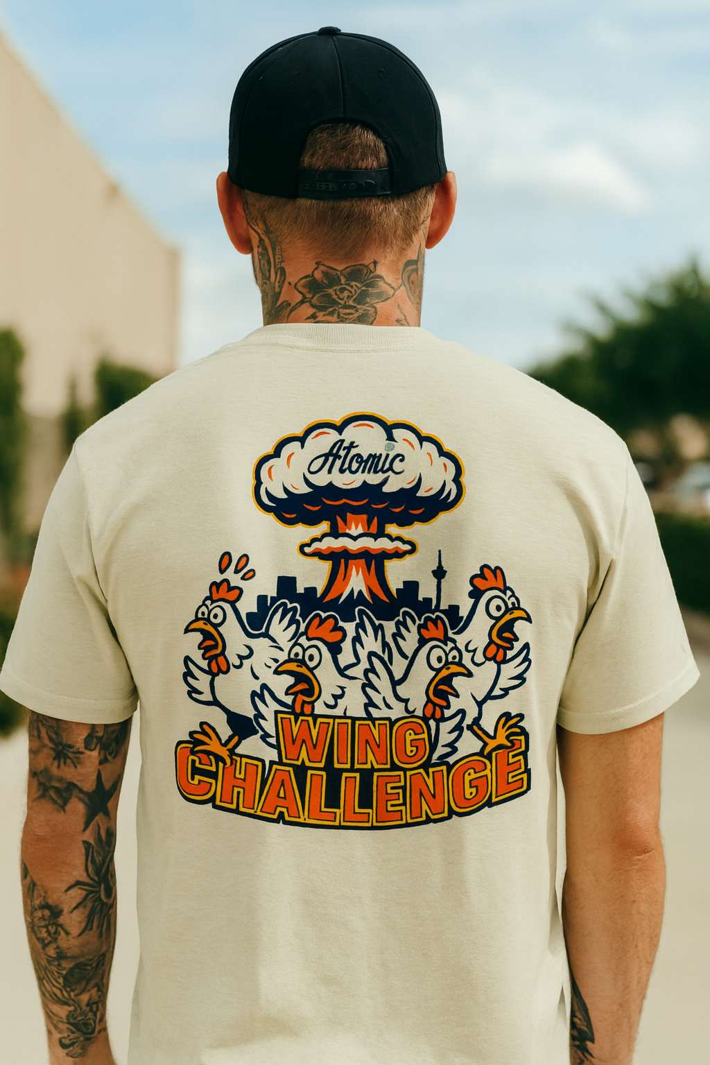Back of a man wearing a black cap and a cream-colored T-shirt with a graphic design featuring chickens, a mushroom cloud, and the words 'Atomic Wing Challenge'.