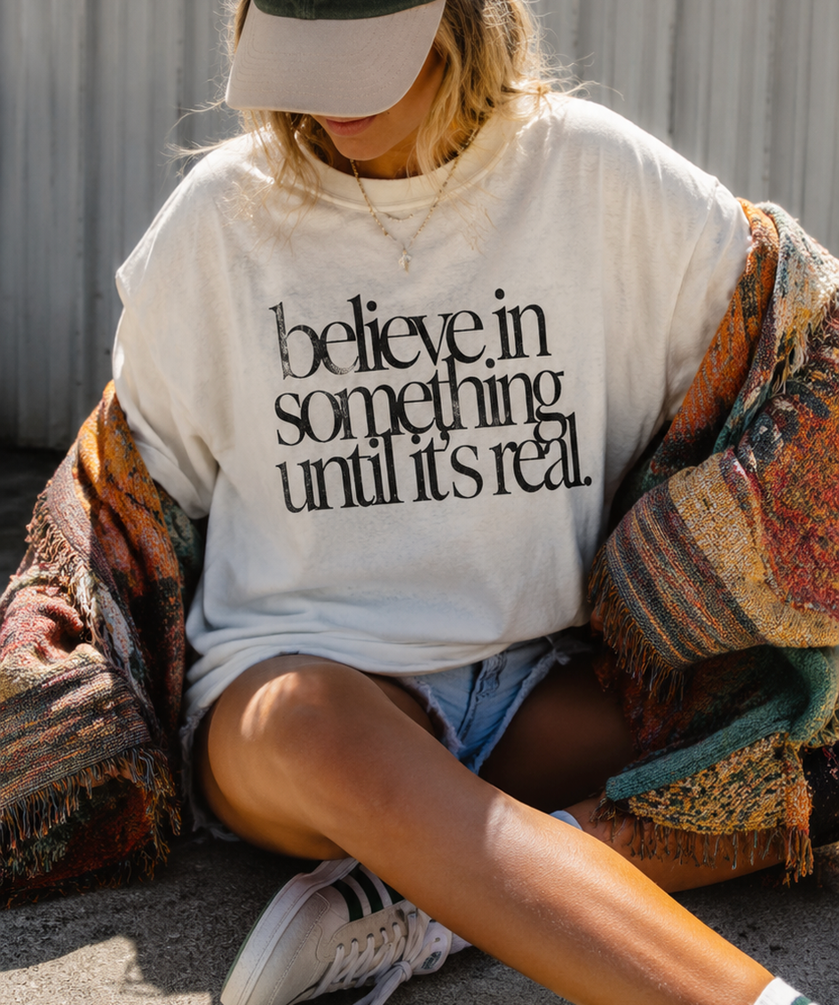Person sitting outdoors wearing a white t-shirt with the text 'believe in something until it's real', denim shorts, a colorful woven blanket, white sneakers, a beige cap, and layered necklaces.