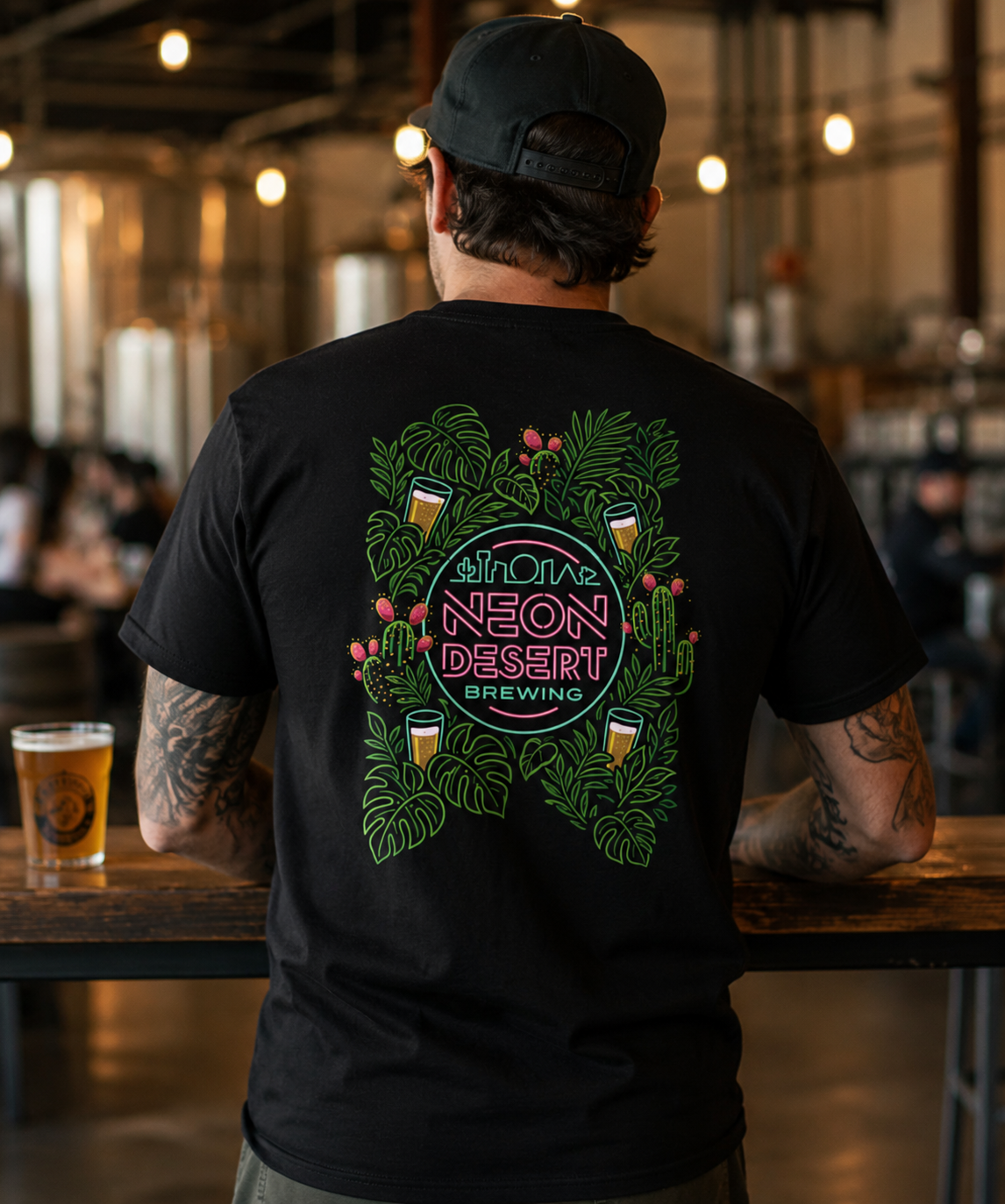 Back of a person wearing a black T-shirt with neon-colored jungle-themed graphic and text promoting Neon Desert Brewing, inside a brewery or bar setting.