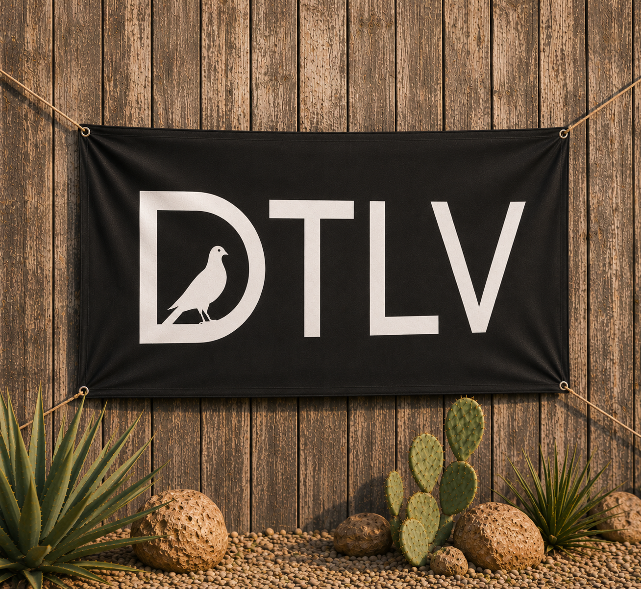 A black banner with the white letters D T I L V, and a pigeon silhouette inside the D, hanging on a wooden fence with desert plants and rocks in front.