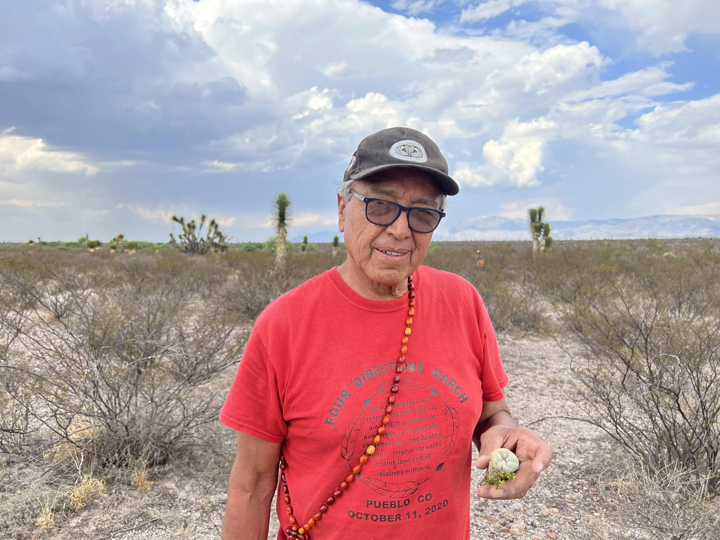 A man wearing glasses, a black cap, and a red T-shirt holding a small plant, standing in a desert landscape with sparse bushes and a cloudy sky.