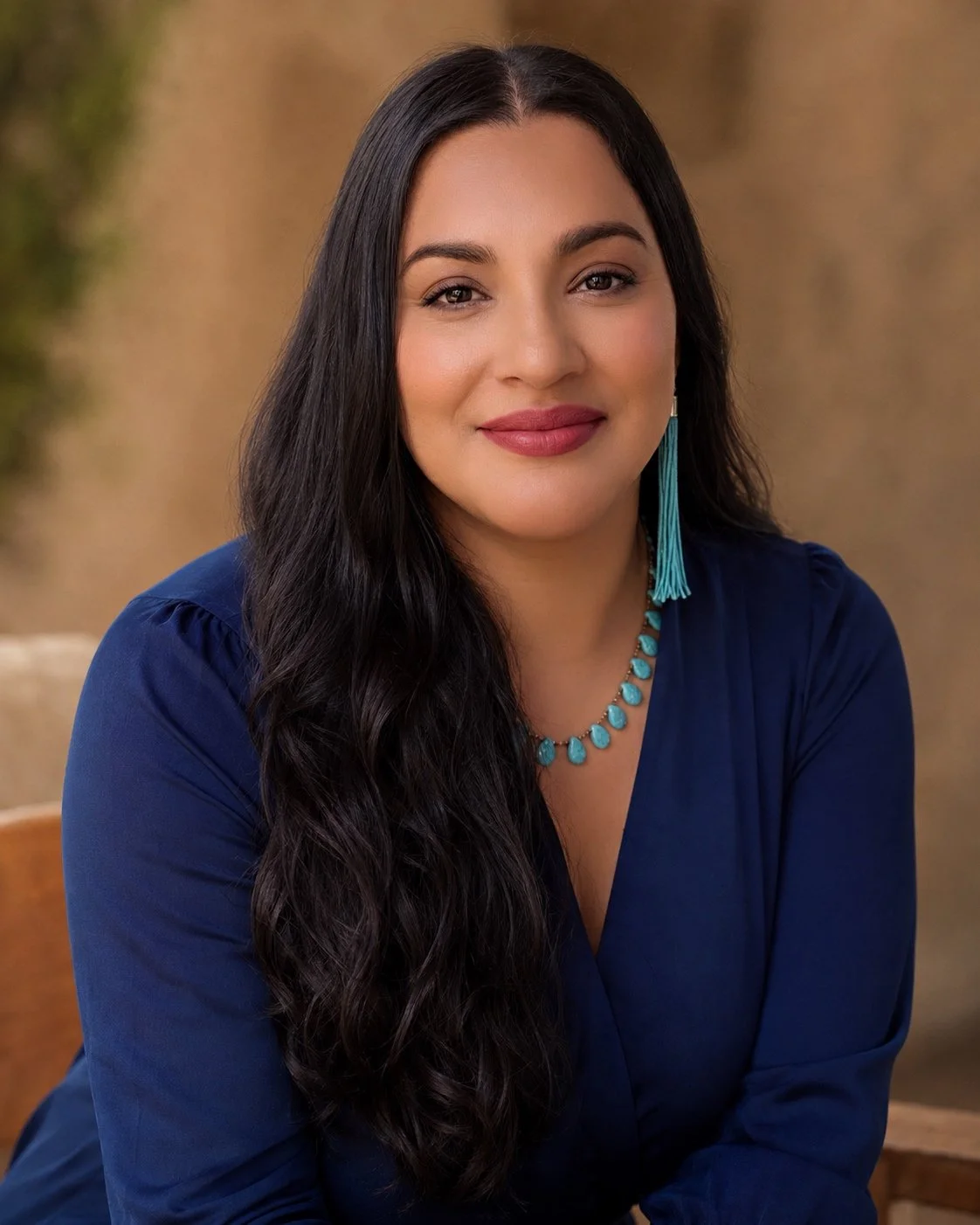 A woman with long dark hair, wearing a blue dress, turquoise jewelry including a necklace, earrings, and a bracelet, sitting outdoors with a blurred natural background.