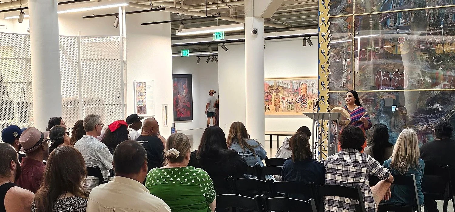 A woman is speaking at a podium in a gallery, giving a presentation to an audience seated and listening. The gallery has artwork on the walls and large metal posts supporting the ceiling.