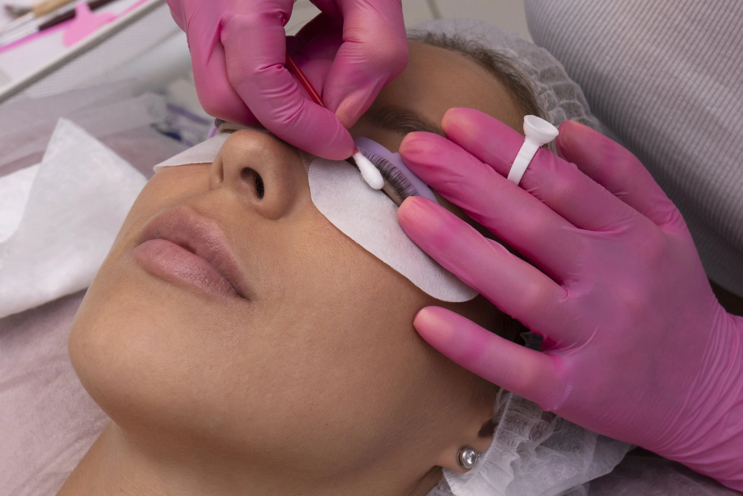 A person is lying down with their eye protected by a medical pad, as a technician in pink gloves applies eyelash extensions.