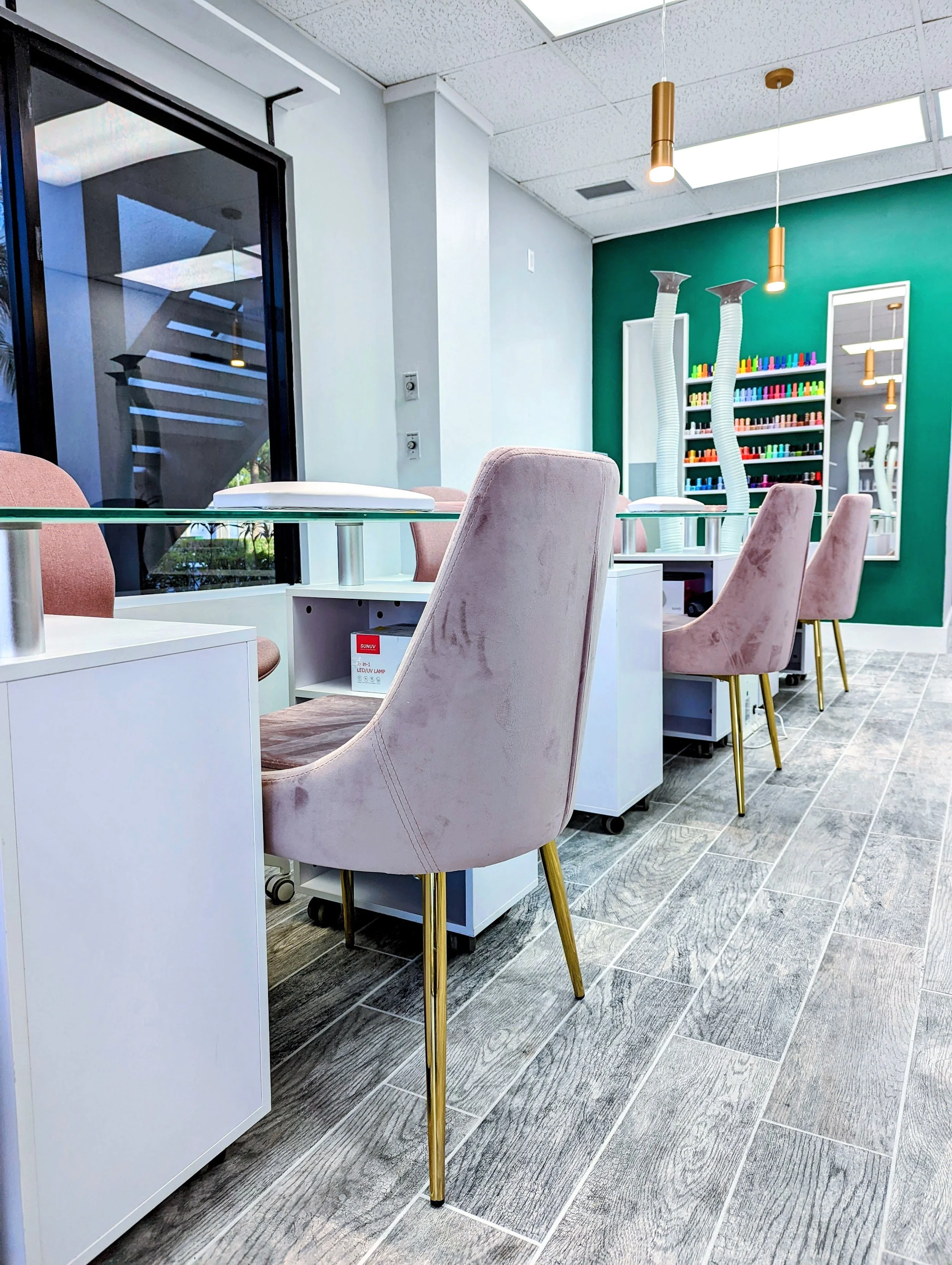 Interior of a nail salon with pastel pink velvet chairs, white nail stations, and a green accent wall with shelves of nail polish bottles.