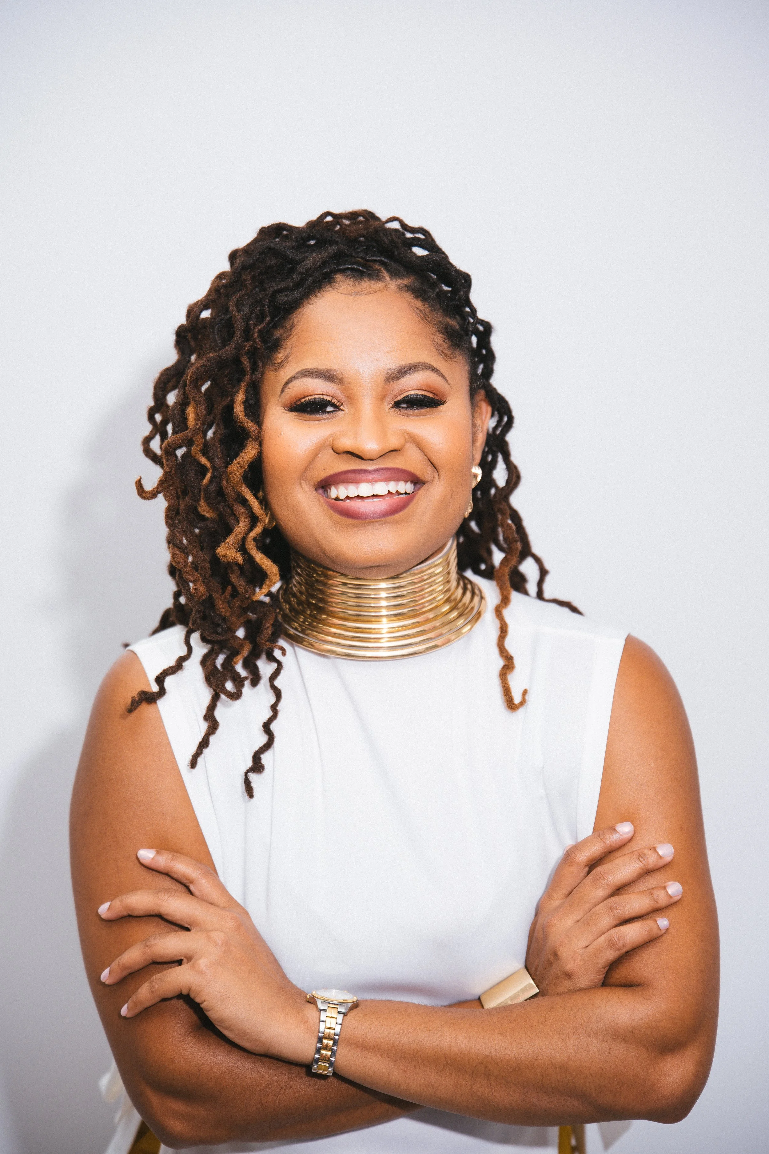 A smiling woman with dark braided hair, wearing a white sleeveless top, a gold choker necklace, a gold bracelet, and a watch, standing against a plain light background.