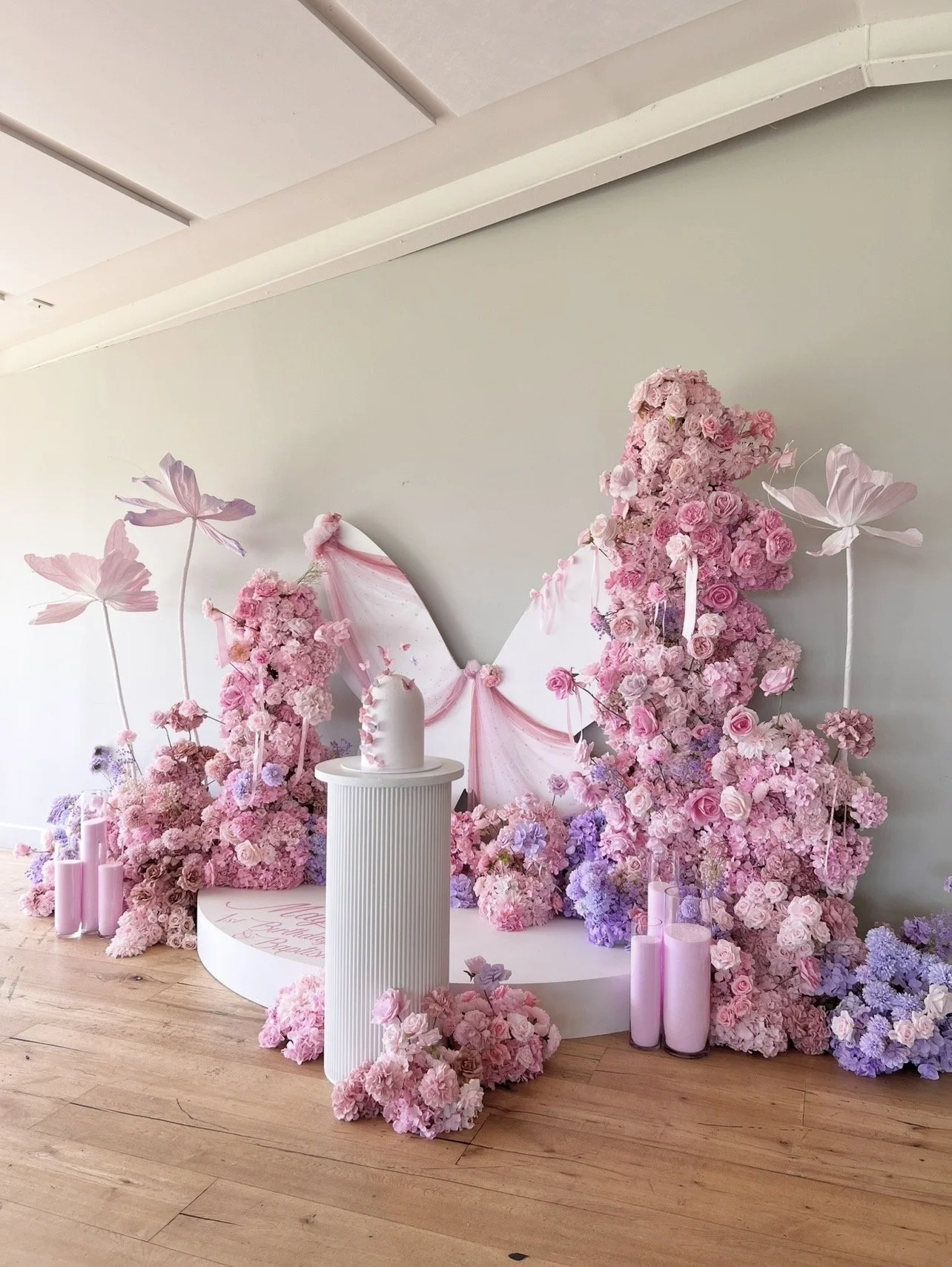 A decorative floral display with pink and purple flowers, candles, and pastel pink drapery, likely for a celebration or event, set against a light green wall.