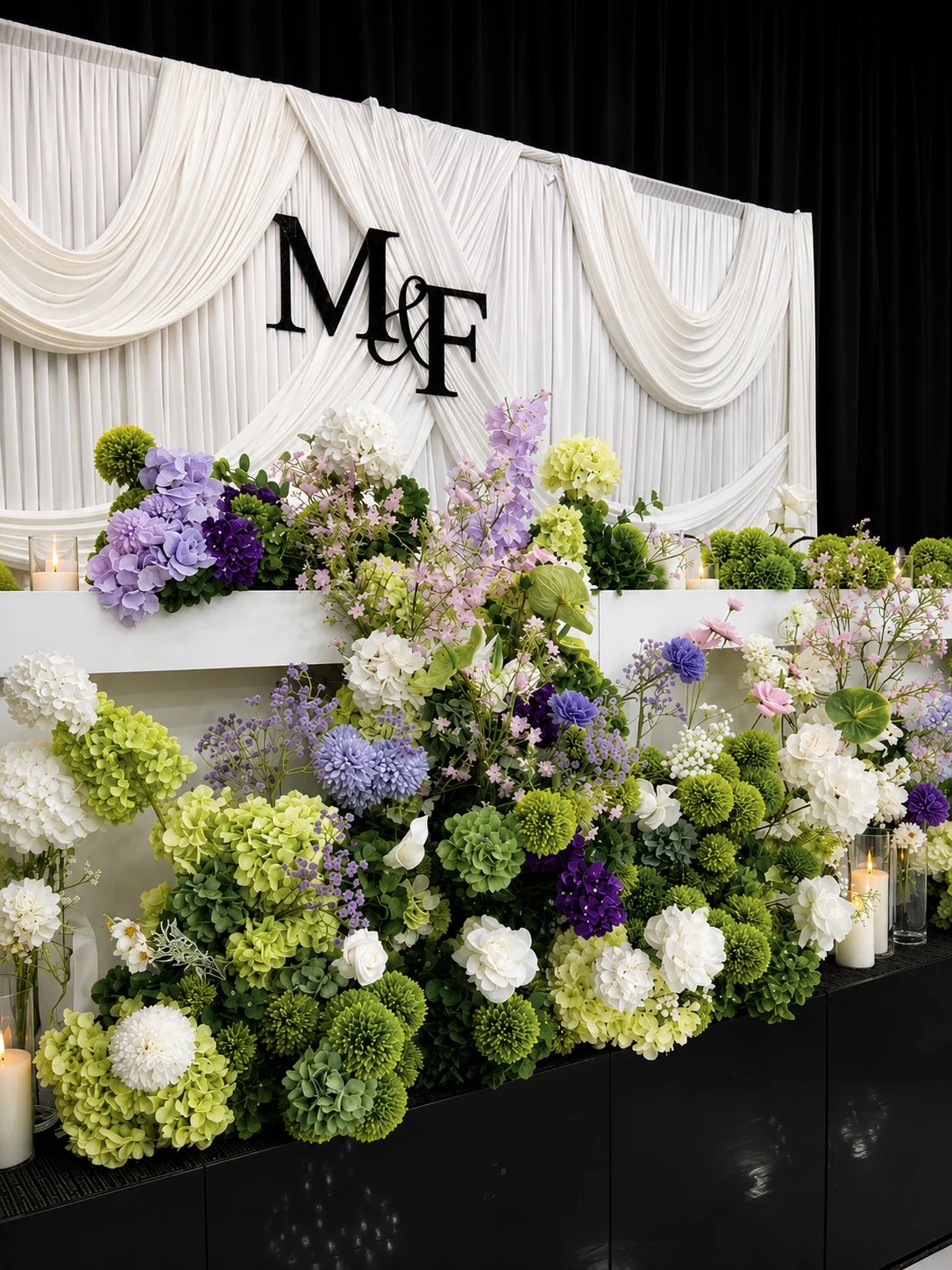 Wedding or event floral arrangement with white, purple, and green flowers in front of a white draped backdrop with black 'M & F' initials and black curtains