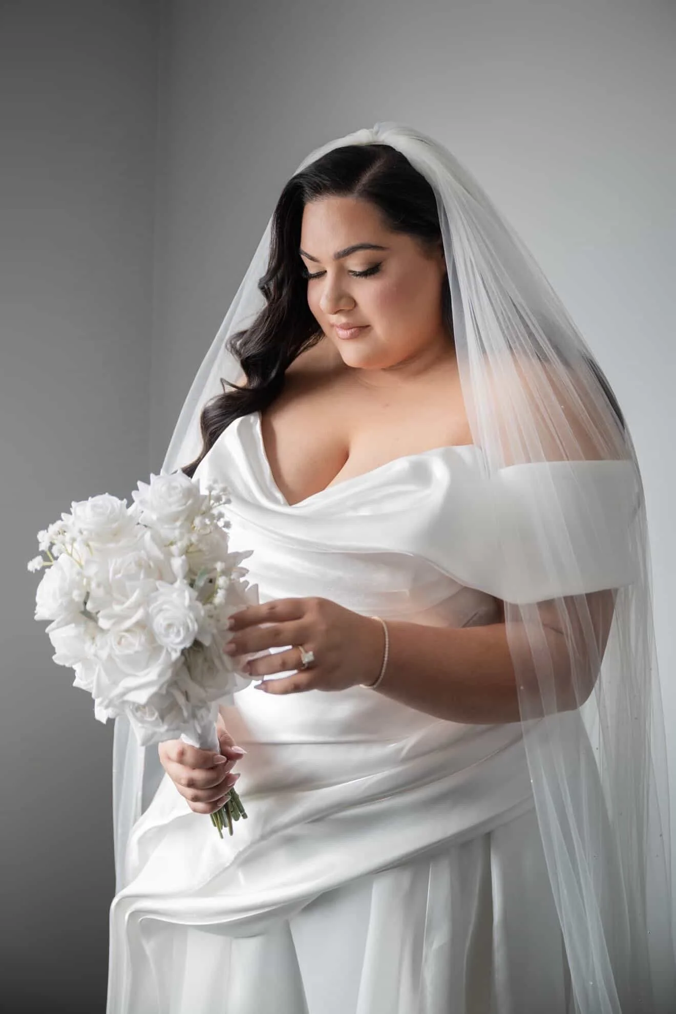 A woman in a white wedding dress holding a bouquet of white roses, with a veil over her dark, wavy hair, looking down with her eyes closed.