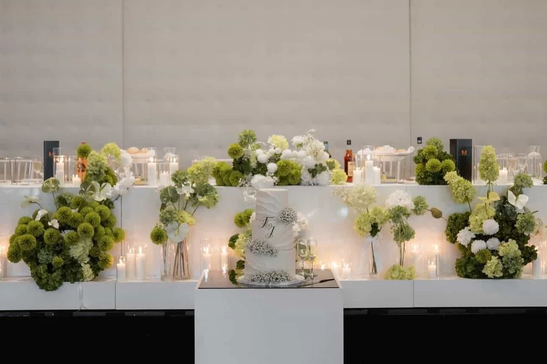 Elegant table with white and green floral arrangements, candles, and a white wedding cake at a celebration or event.