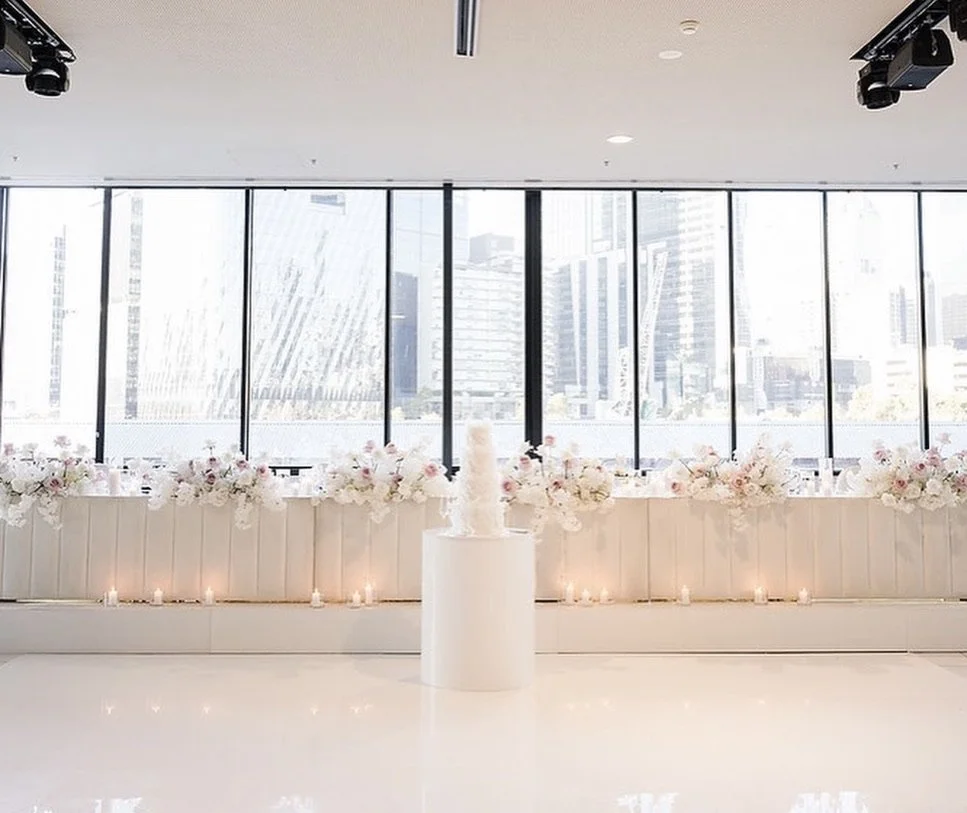 A bright indoor space with a table decorated with white and pink flowers, a tall white candle, and candles in glass holders. Large windows in the background reveal a cityscape with tall buildings.