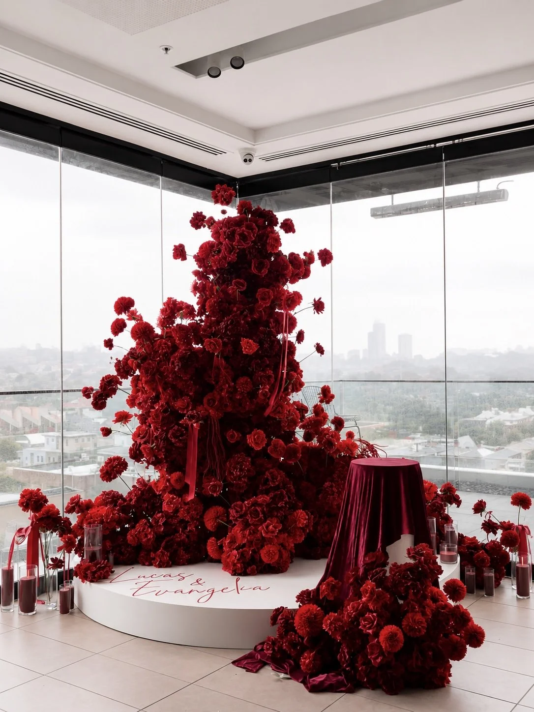 Red floral arrangement sculpture on a white circular platform with a white backdrop, surrounded by red candles and flowers, with cityscape view outside large windows.