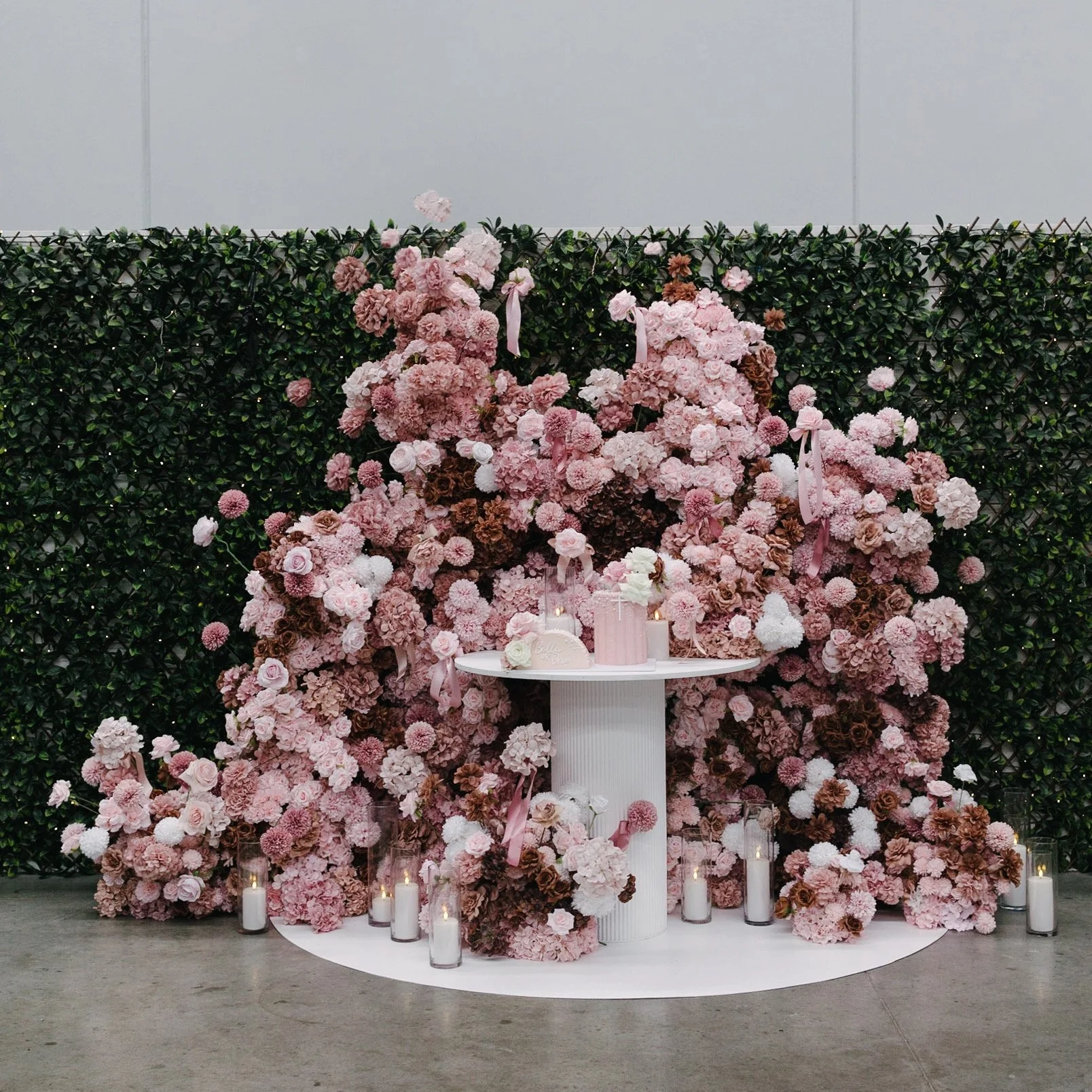 Floral wedding backdrop with pink, white, and brown flowers, candles in glass holders at the base, and a small round table with pink decor, set against a green leafy wall.