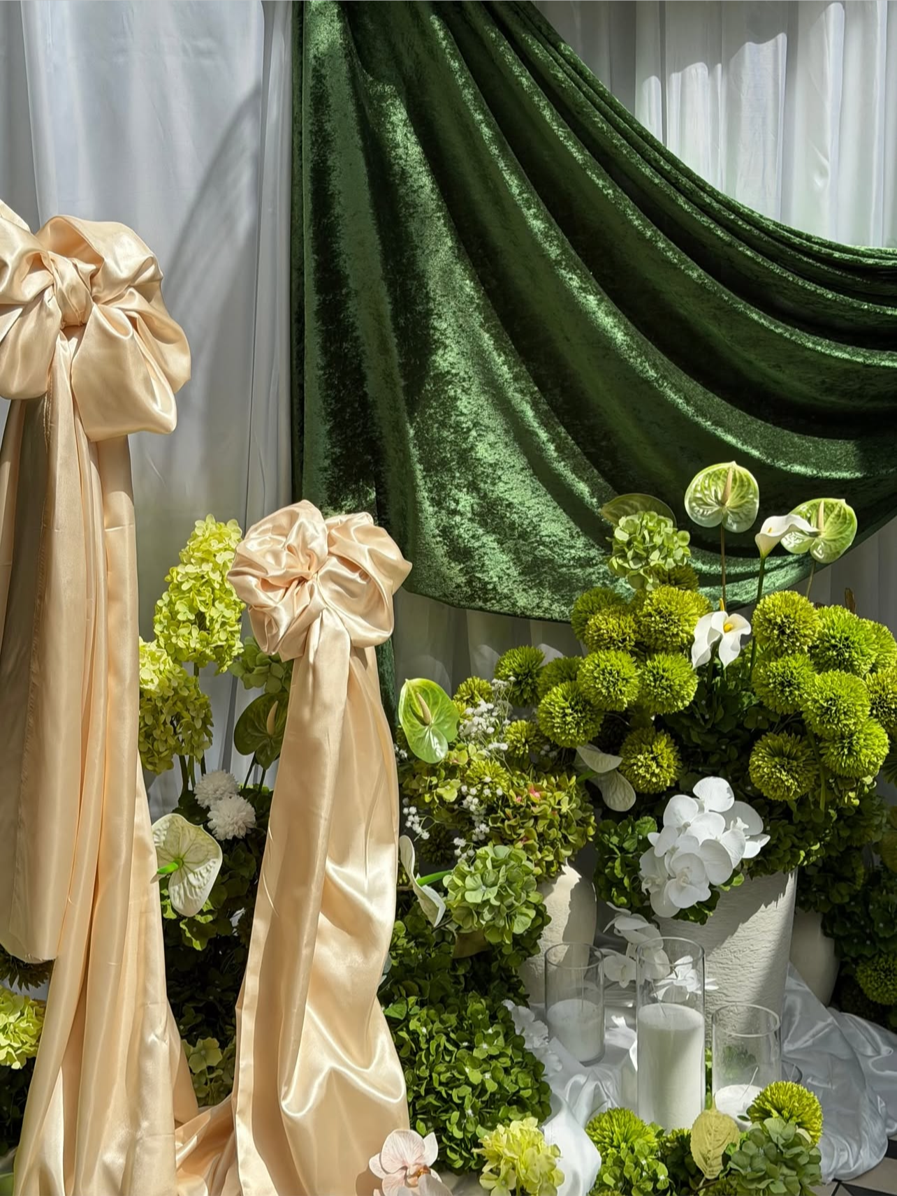 A display of green and white floral arrangements with large cream-colored fabric bows and drapery.