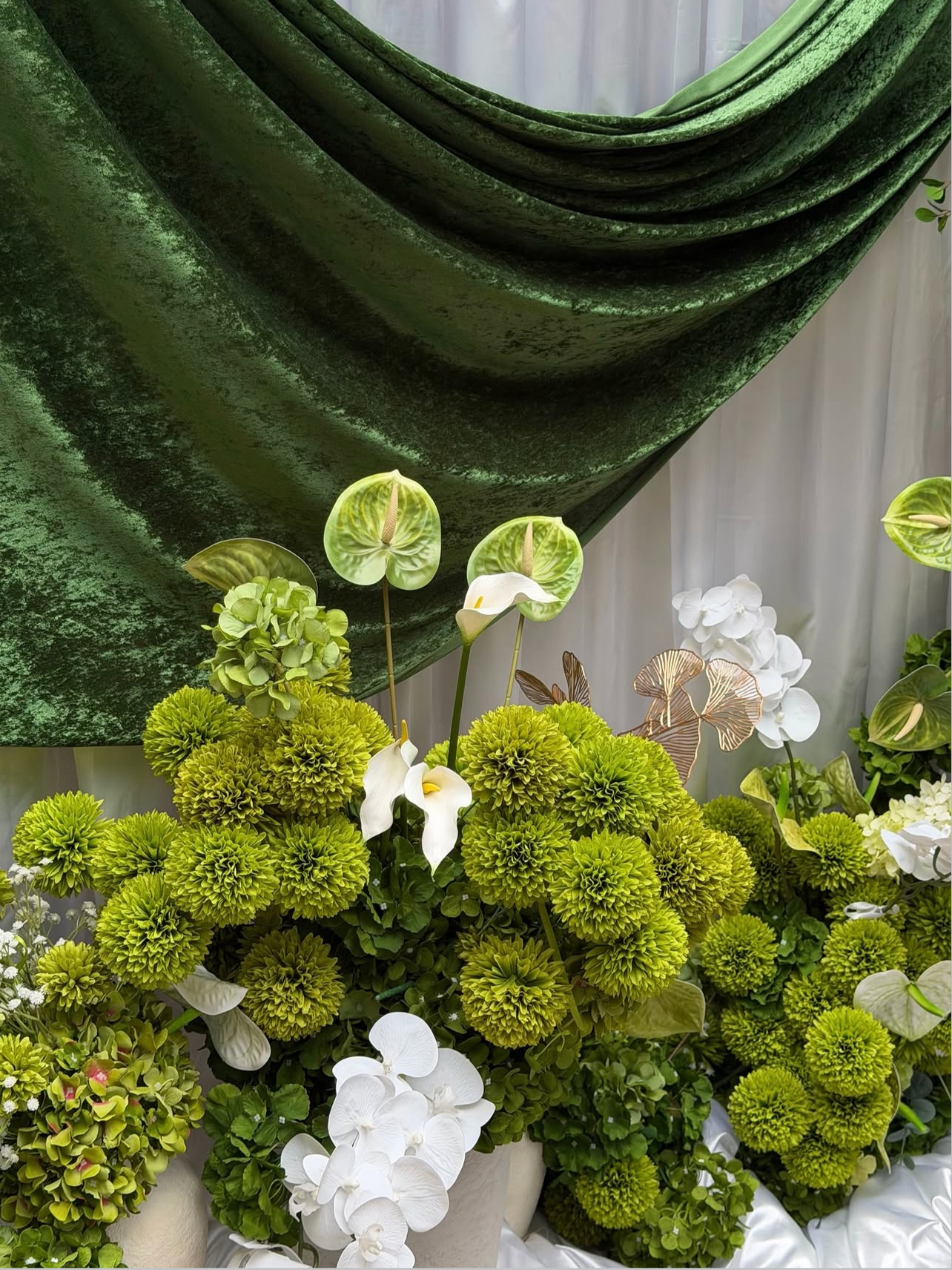 Arrangement of green, white, and cream-colored flowers, including calla lilies, hydrangeas, and mossy green plants, with a large dark green velvet drape in the background.